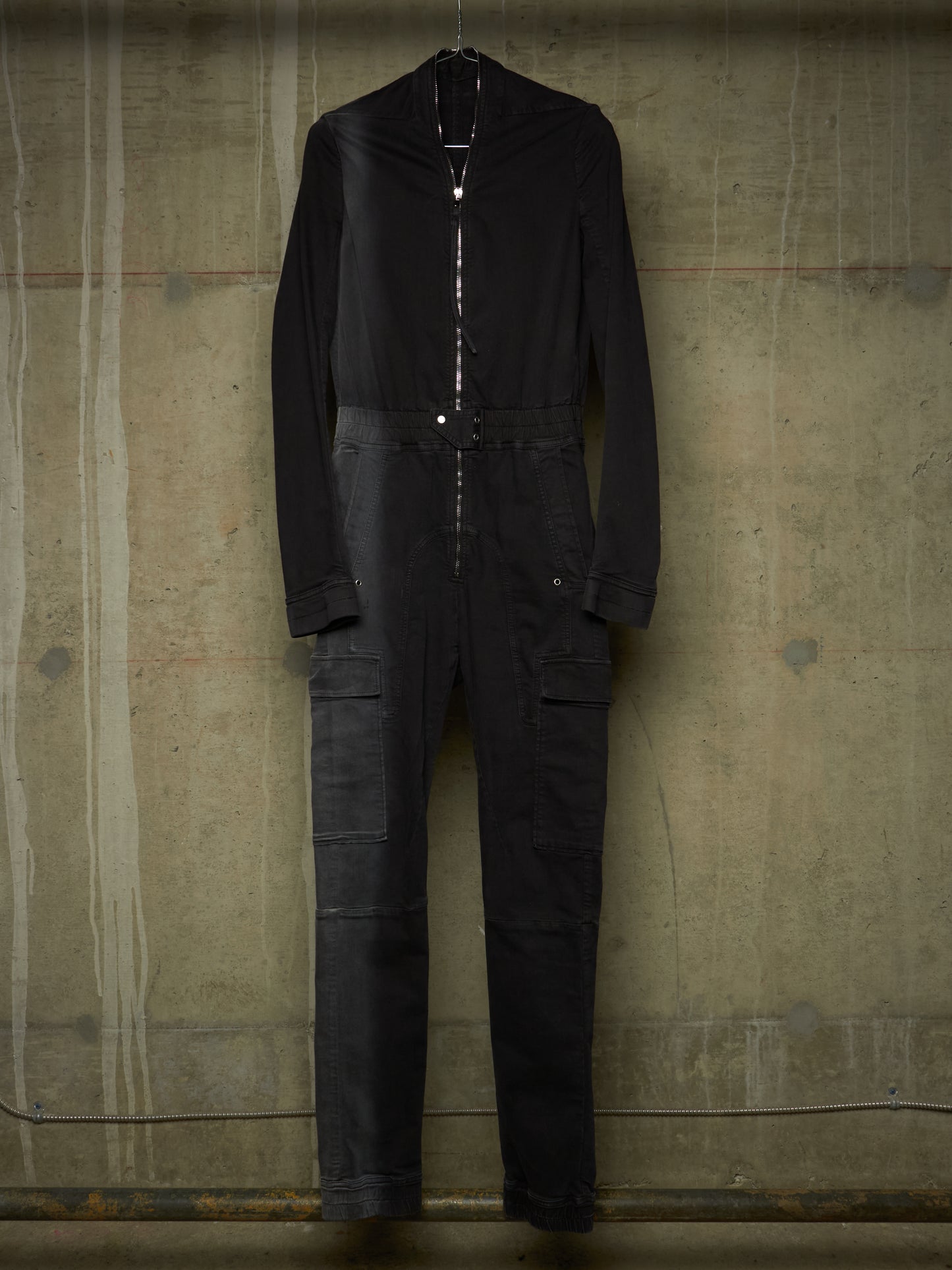 A black long-sleeve jumpsuit with a high collar and front zipper, hanging against a textured concrete wall.