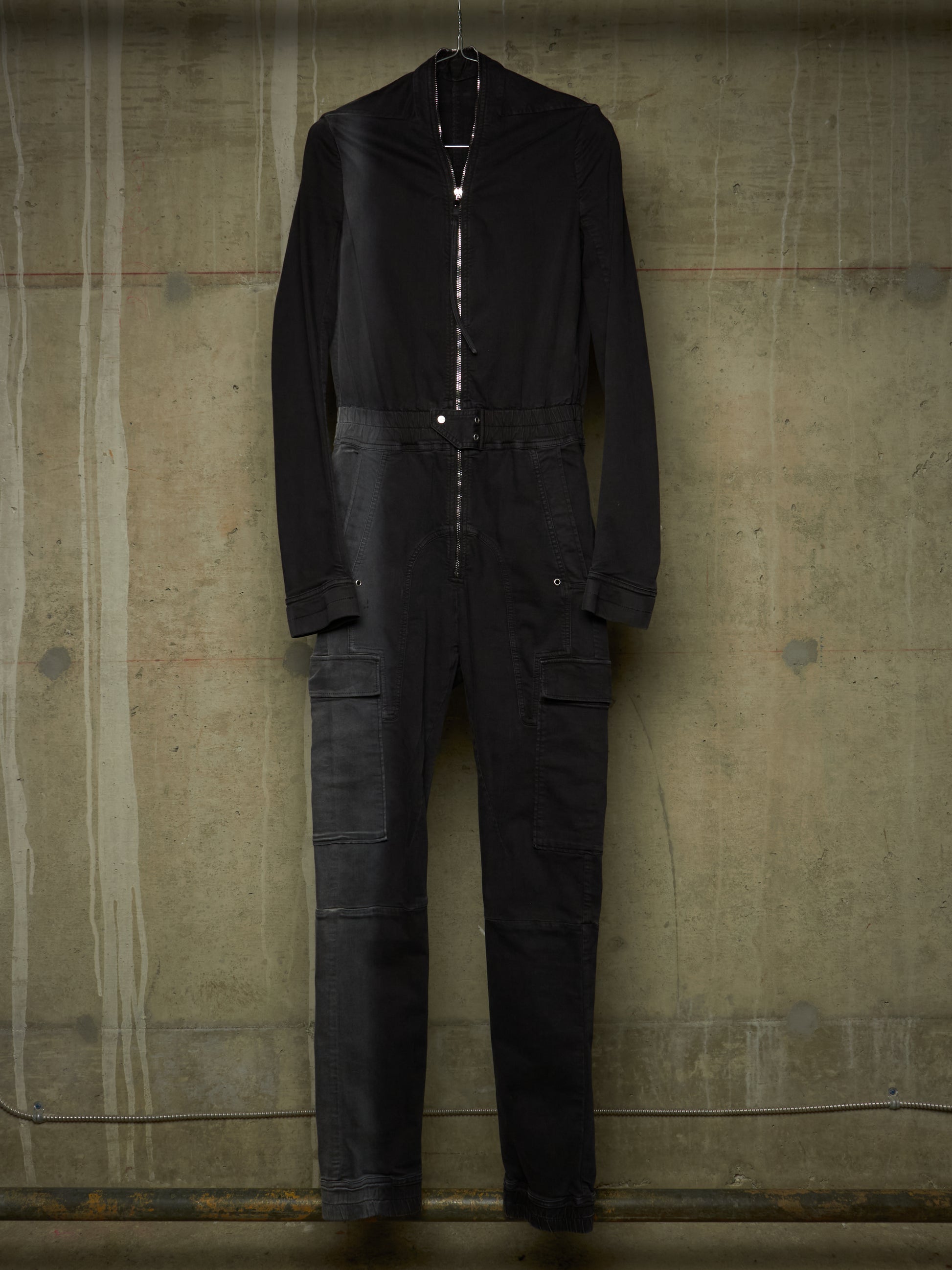 A black long-sleeve jumpsuit with a high collar and front zipper, hanging against a textured concrete wall.