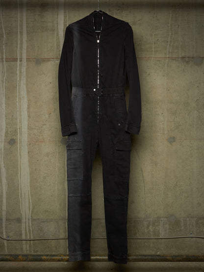 A black long-sleeve jumpsuit with a high collar and front zipper, hanging against a textured concrete wall.