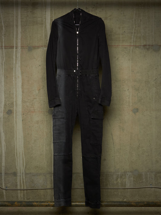 A black long-sleeve jumpsuit with a high collar and front zipper, hanging against a textured concrete wall.