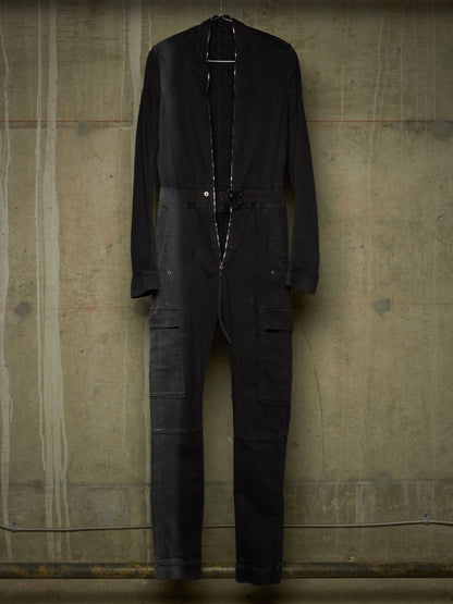 A black long-sleeve utility jumpsuit with a high collar, front zipper, drawstring waist, and cargo pockets, hanging against a textured concrete wall.