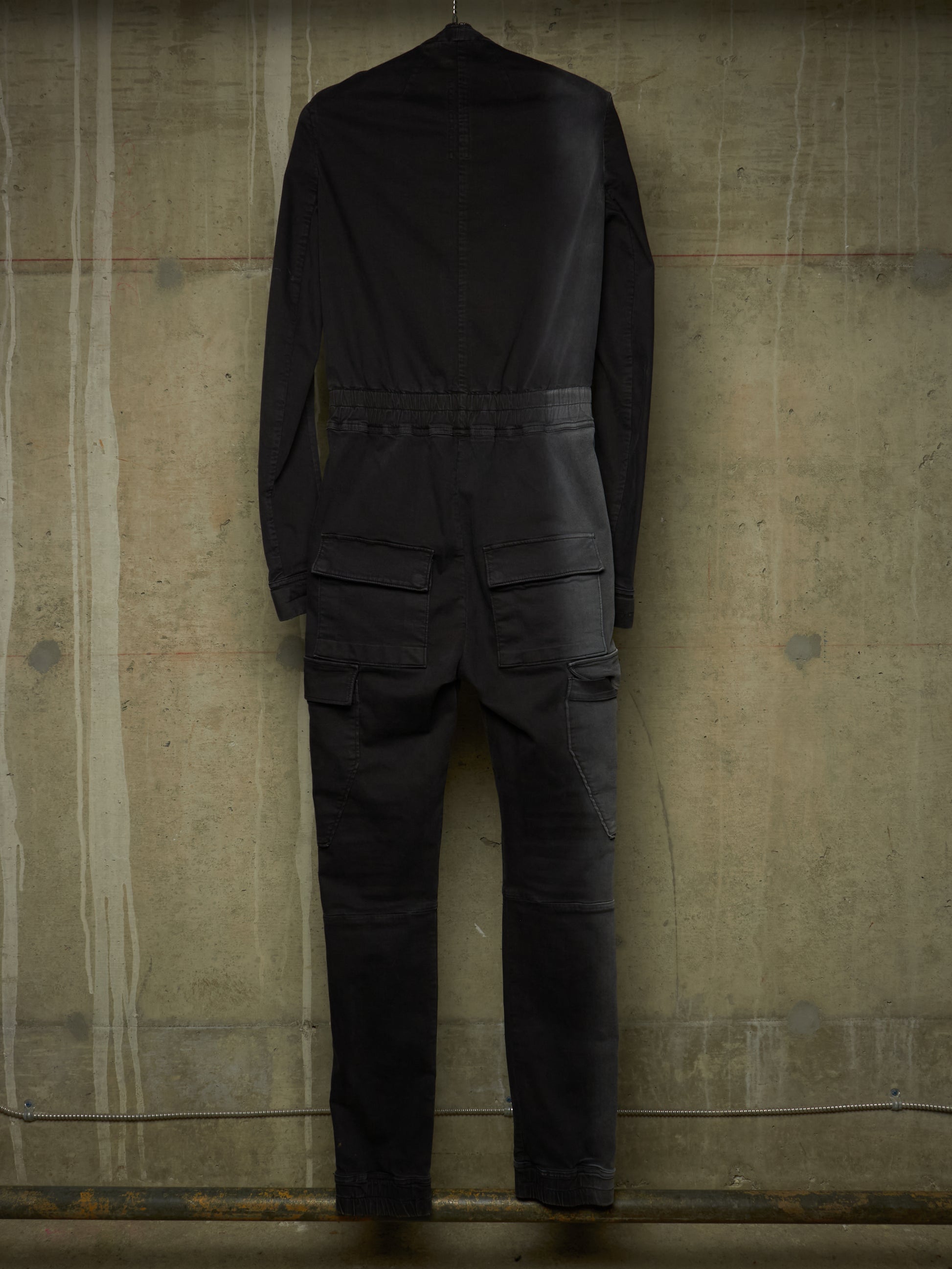 A black long-sleeve jumpsuit or coverall shown from the back, hanging against a textured concrete wall.
