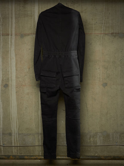 A black long-sleeve jumpsuit or coverall shown from the back, hanging against a textured concrete wall.