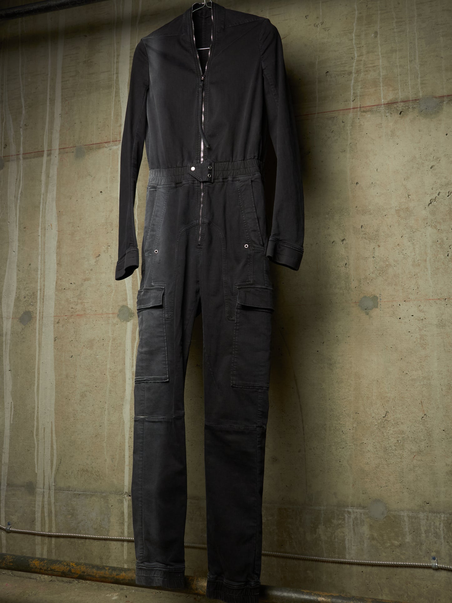 A black long-sleeve utility jumpsuit with a front zipper, elastic waist, and cargo pockets, hanging against a textured concrete wall.