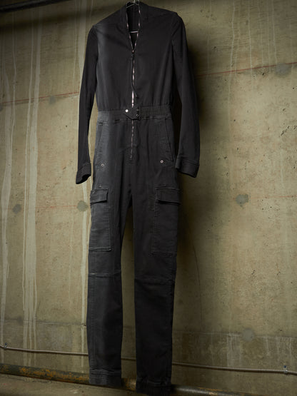 A black long-sleeve utility jumpsuit with a front zipper, elastic waist, and cargo pockets, hanging against a textured concrete wall.