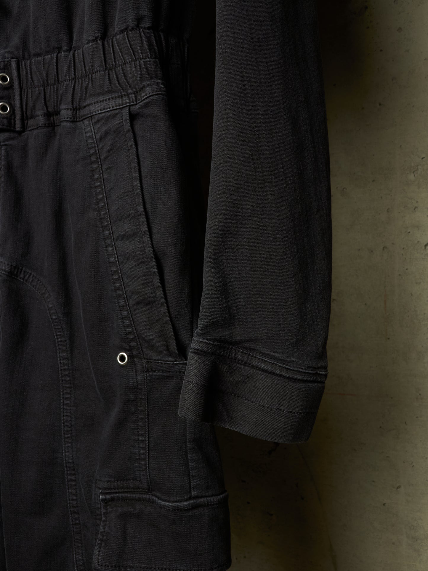 Close-up of a dark charcoal gray or black utility jumpsuit, showing the elastic waistband, side pockets, and rolled cuff of the sleeve against a textured concrete wall.