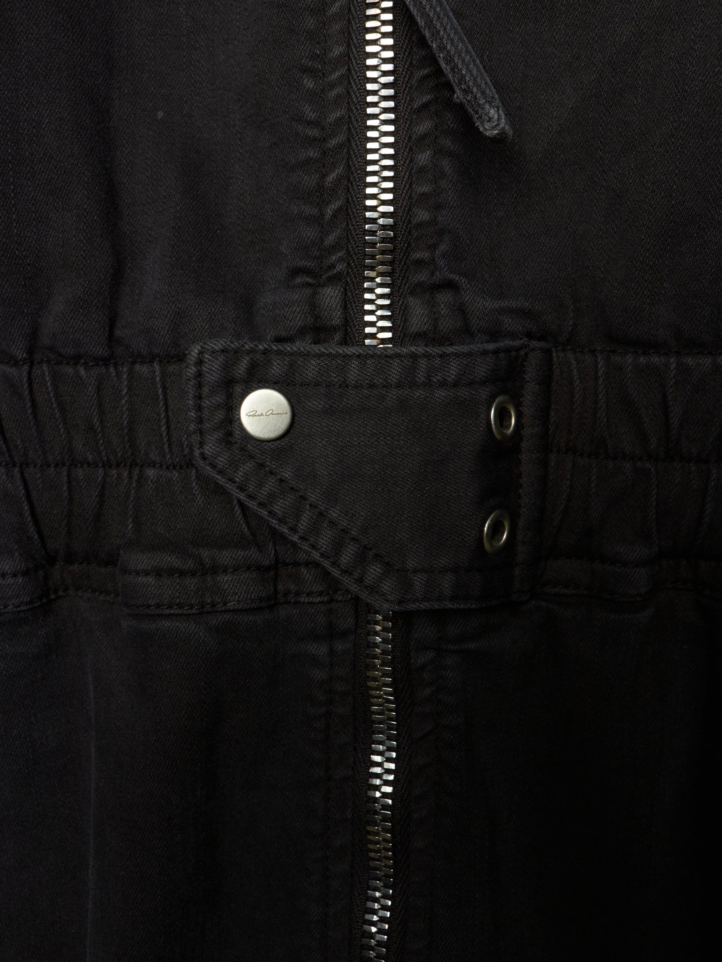 Close-up of a black jacket or overshirt showing the waist area with a central zipper, a flap pocket with a silver snap button, and two metal eyelets.