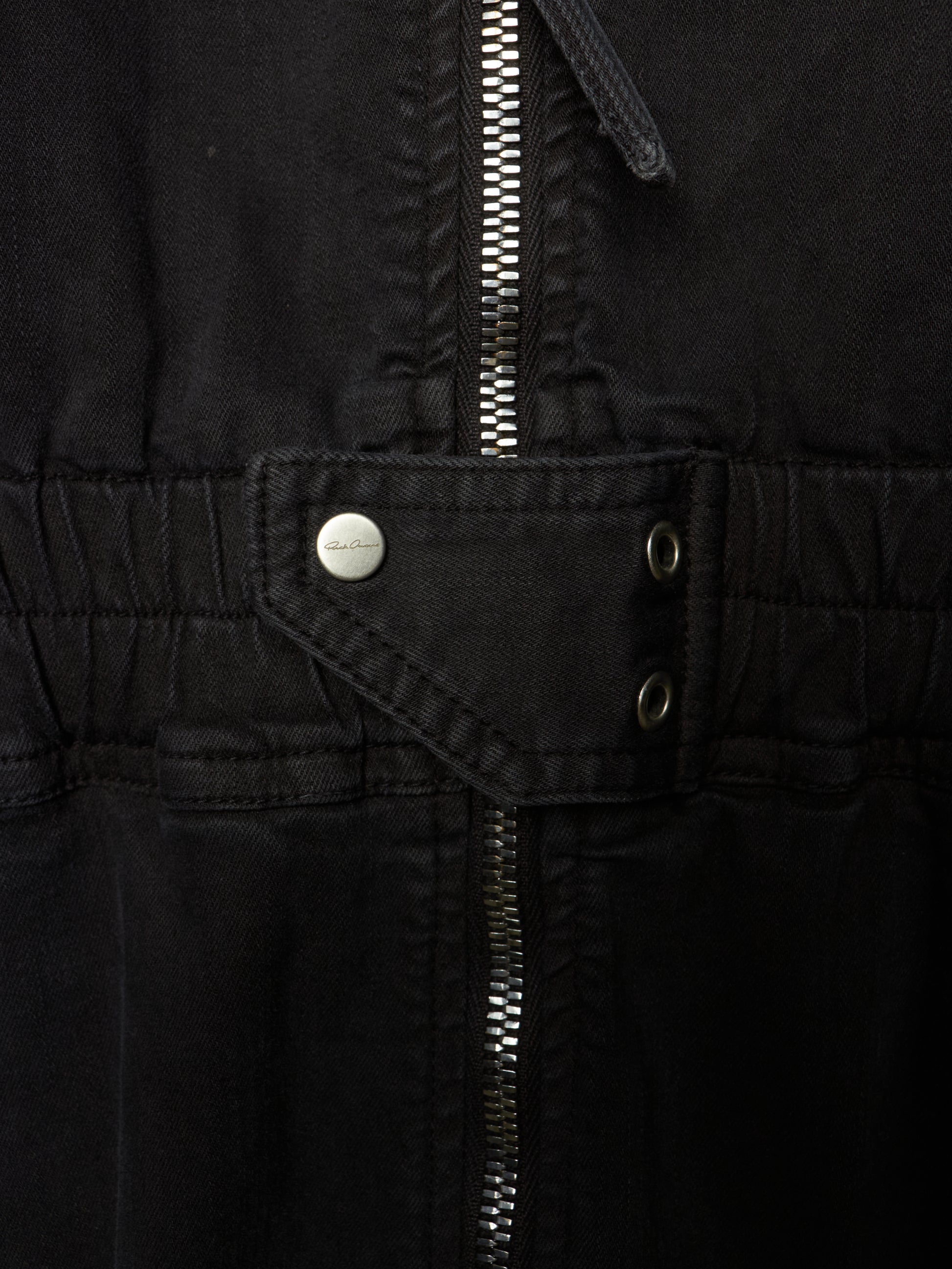 Close-up of a black jacket or overshirt showing the waist area with a central zipper, a flap pocket with a silver snap button, and two metal eyelets.