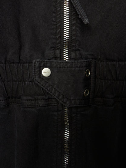 Close-up of a black jacket or overshirt showing the waist area with a central zipper, a flap pocket with a silver snap button, and two metal eyelets.