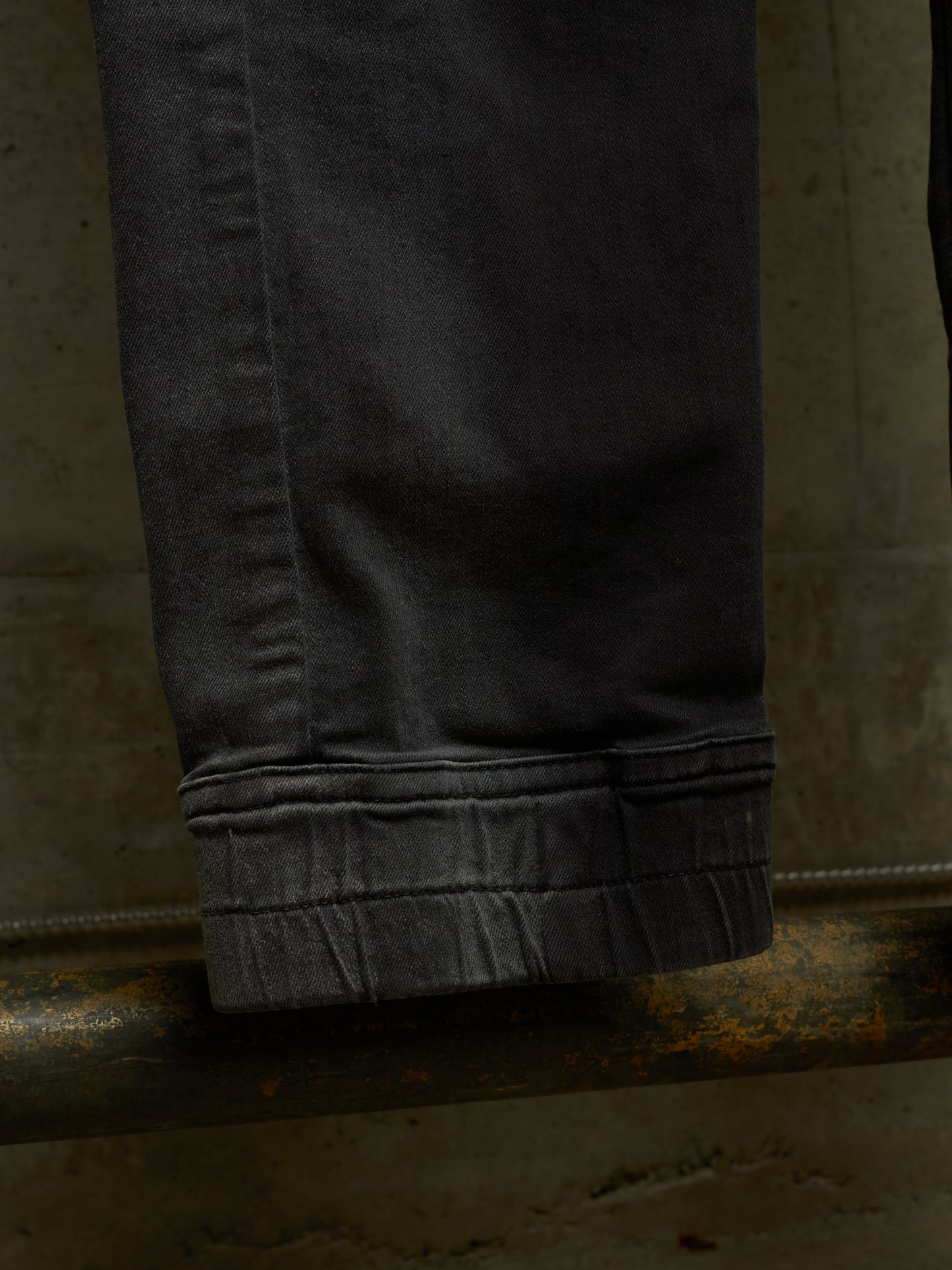 Close-up of a dark charcoal gray pair of jeans folded over to show the folded cuff, resting on a rusty metal pipe against a textured concrete wall.