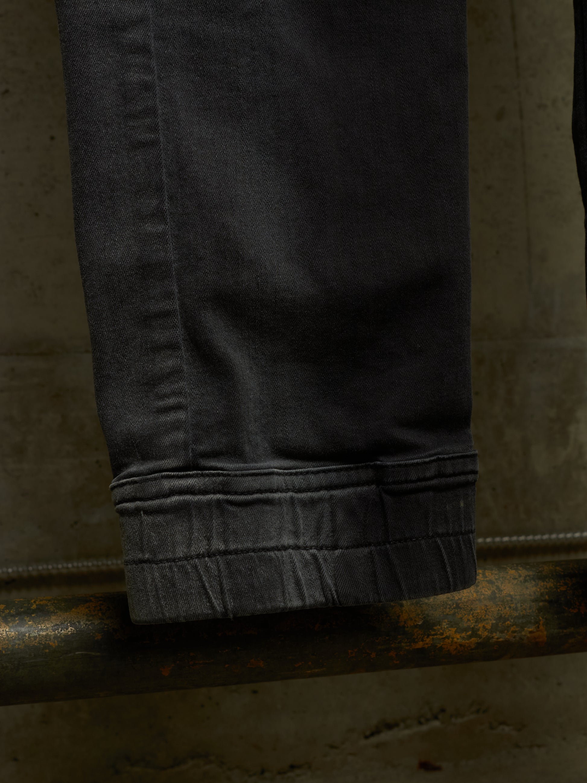 Close-up of a dark charcoal gray pair of jeans folded over to show the folded cuff, resting on a rusty metal pipe against a textured concrete wall.
