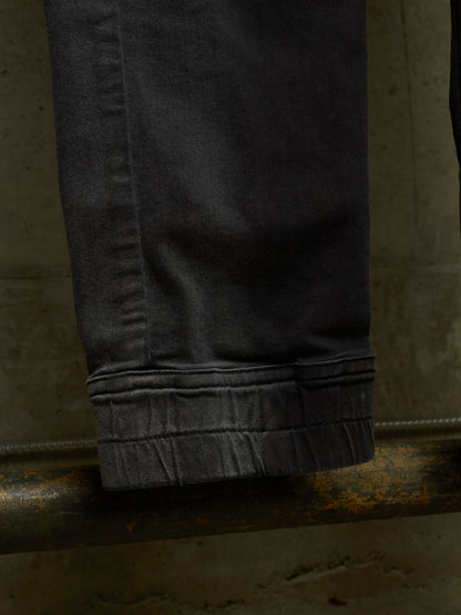 Close-up of a dark charcoal gray pair of jeans folded over to show the folded cuff, resting on a rusty metal pipe against a textured concrete wall.