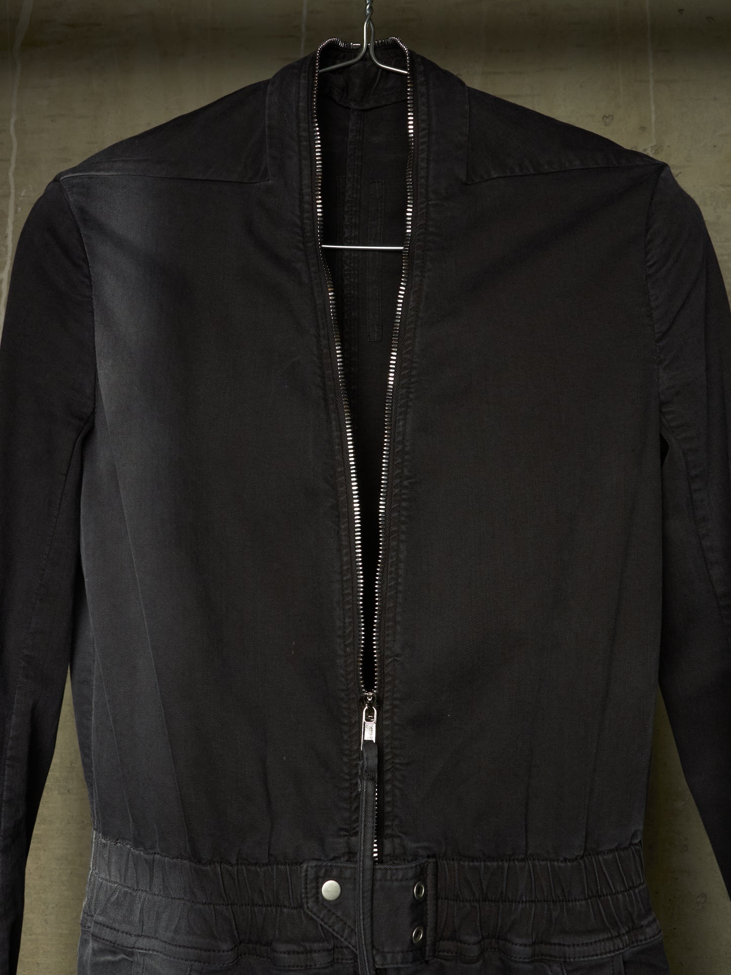 A black zip-up jacket hanging on a metal hanger against a textured concrete wall.