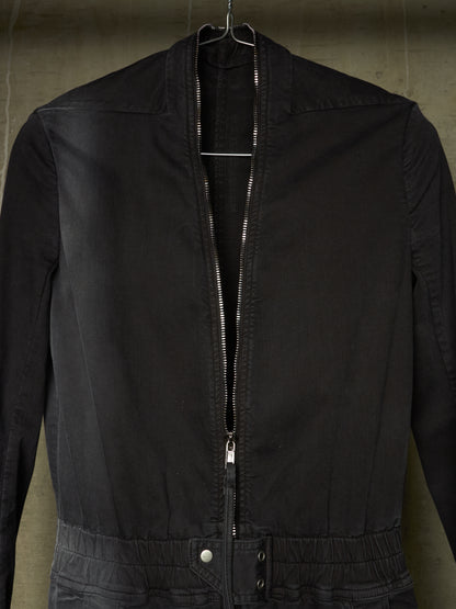A black zip-up jacket hanging on a metal hanger against a textured concrete wall.