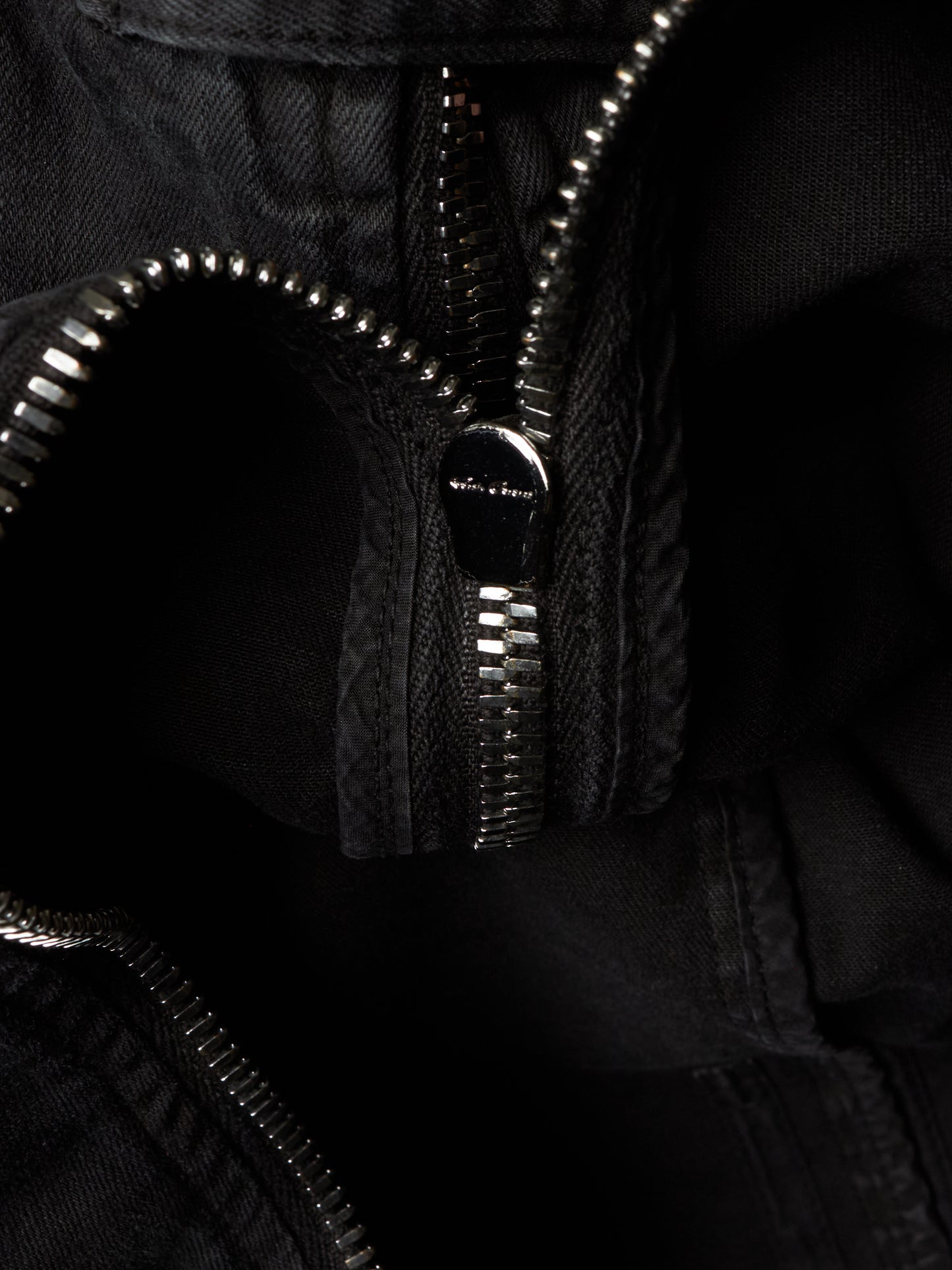 Close-up detail of a black zippered garment, showing the silver-toned metal zipper teeth and a small metallic brand plaque on the zipper pull.