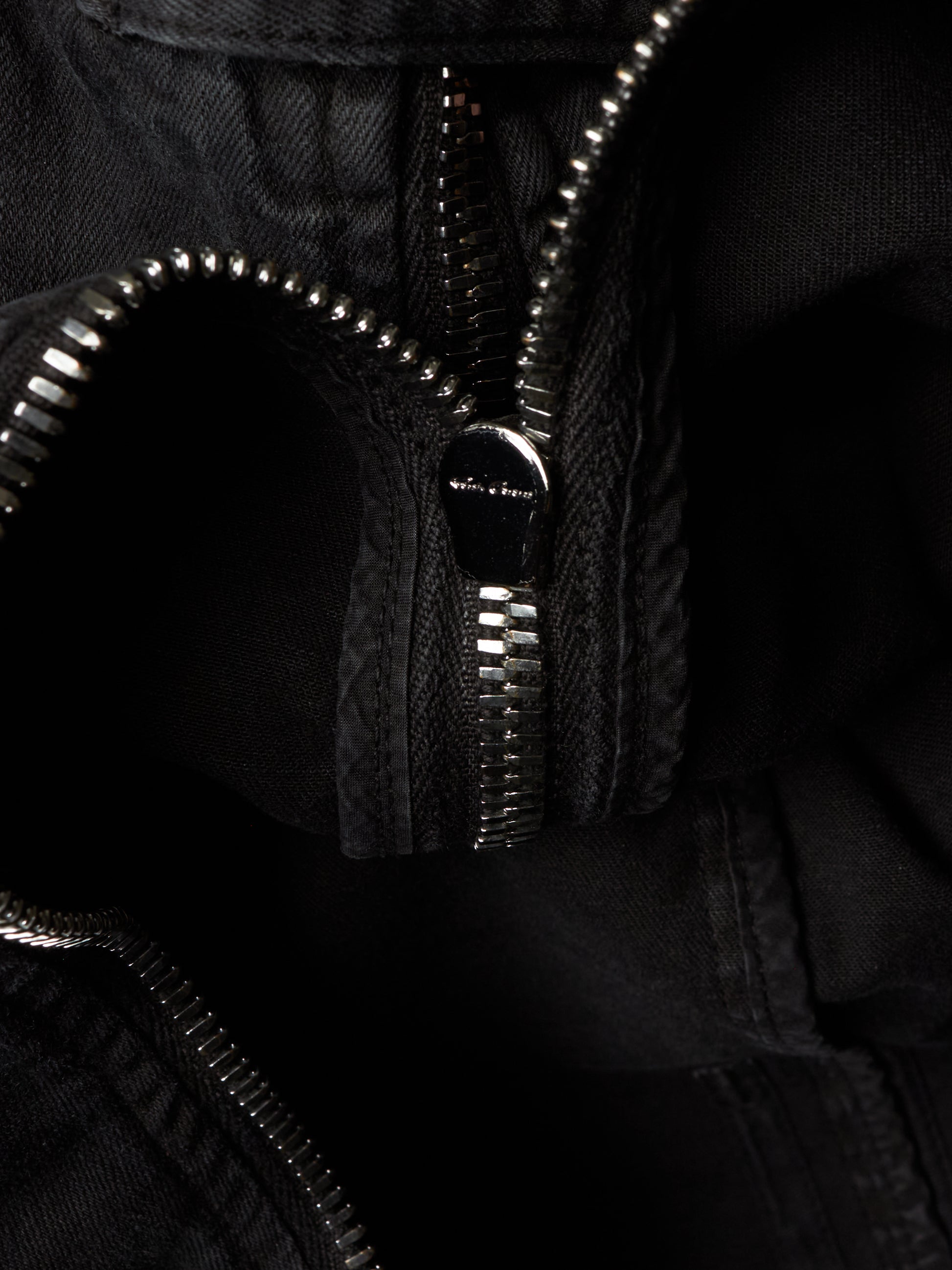 Close-up detail of a black zippered garment, showing the silver-toned metal zipper teeth and a small metallic brand plaque on the zipper pull.