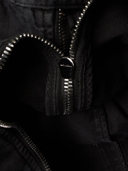 Close-up detail of a black zippered garment, showing the silver-toned metal zipper teeth and a small metallic brand plaque on the zipper pull.