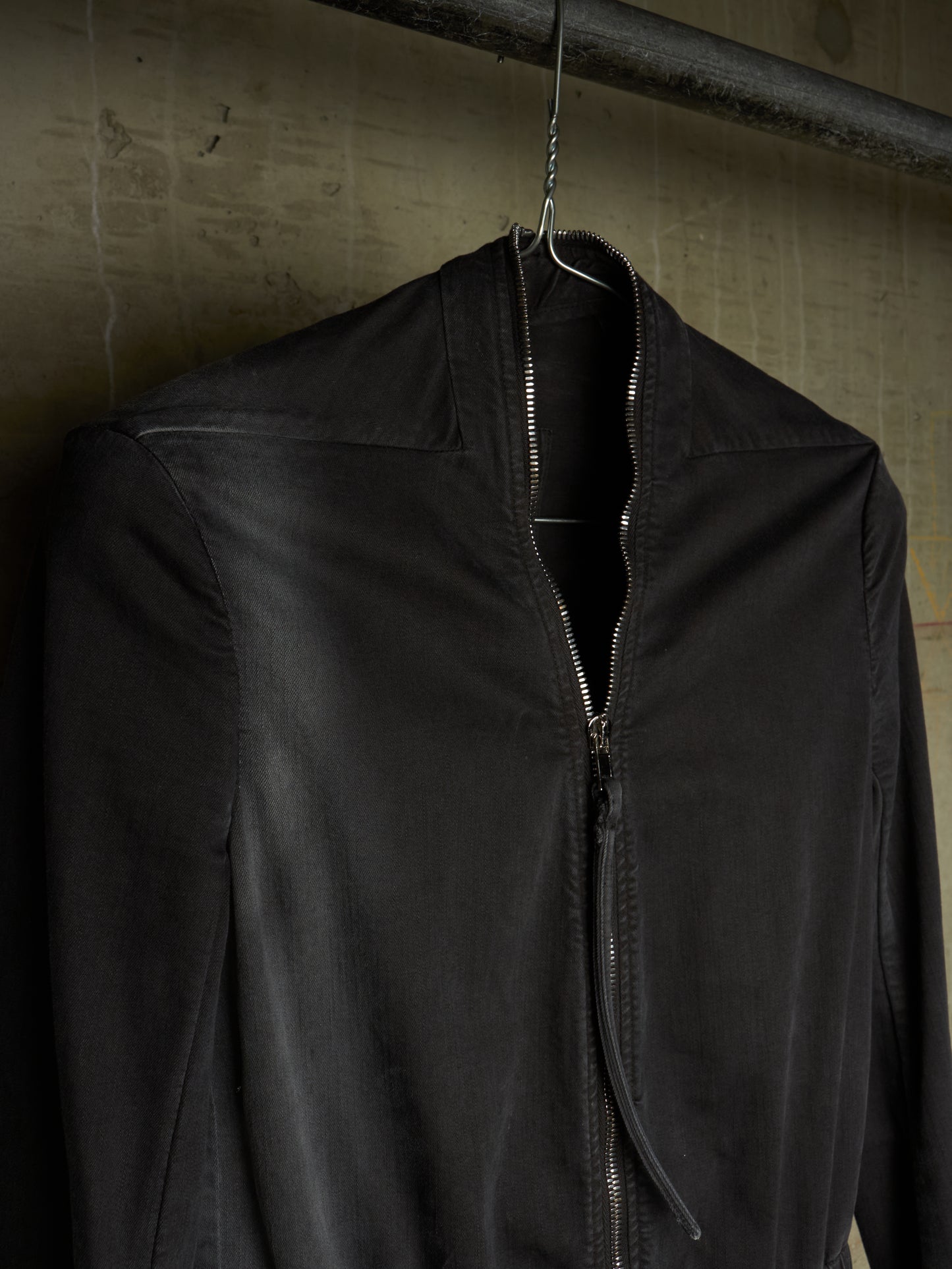 A black zip-up jacket hanging on a metal hanger against a textured concrete wall.