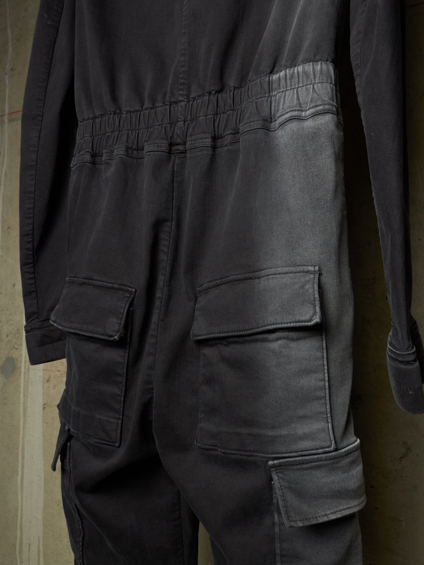 Close-up of the back of a dark charcoal gray jumpsuit or coverall with an elastic waistband, large flap cargo pockets, and long sleeves, shown against a muted brown background.