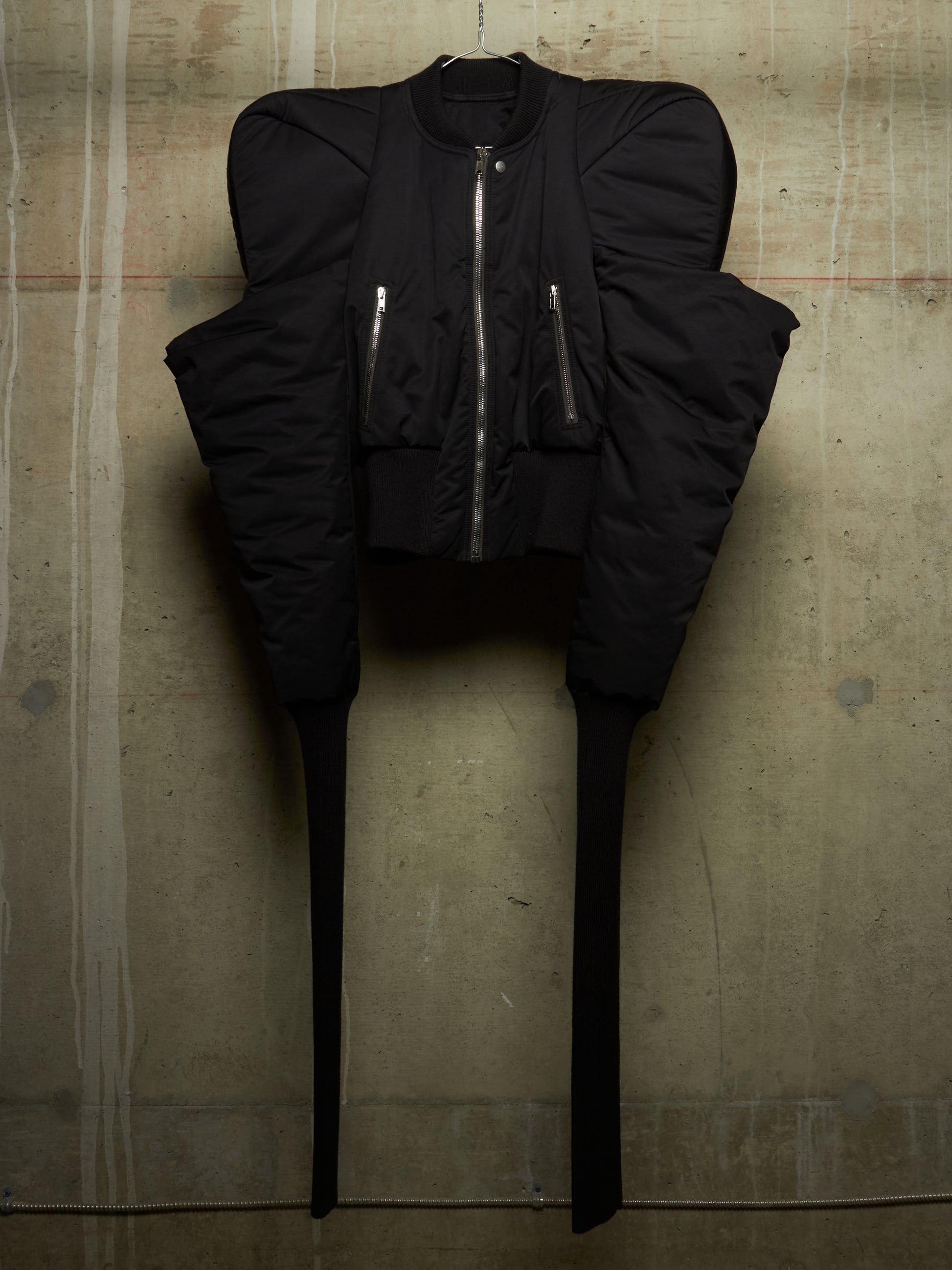 A black futuristic puffer jacket with dramatic shoulder flares, a front zipper, and long narrow legs, hanging against a textured concrete wall.