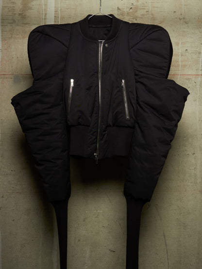 A black puffer jacket with dramatic, oversized structured shoulders and long tapering legs, hanging against a textured concrete wall. The jacket features a central silver zipper, two vertical zippered chest pockets, and ribbed cuffs and hem.