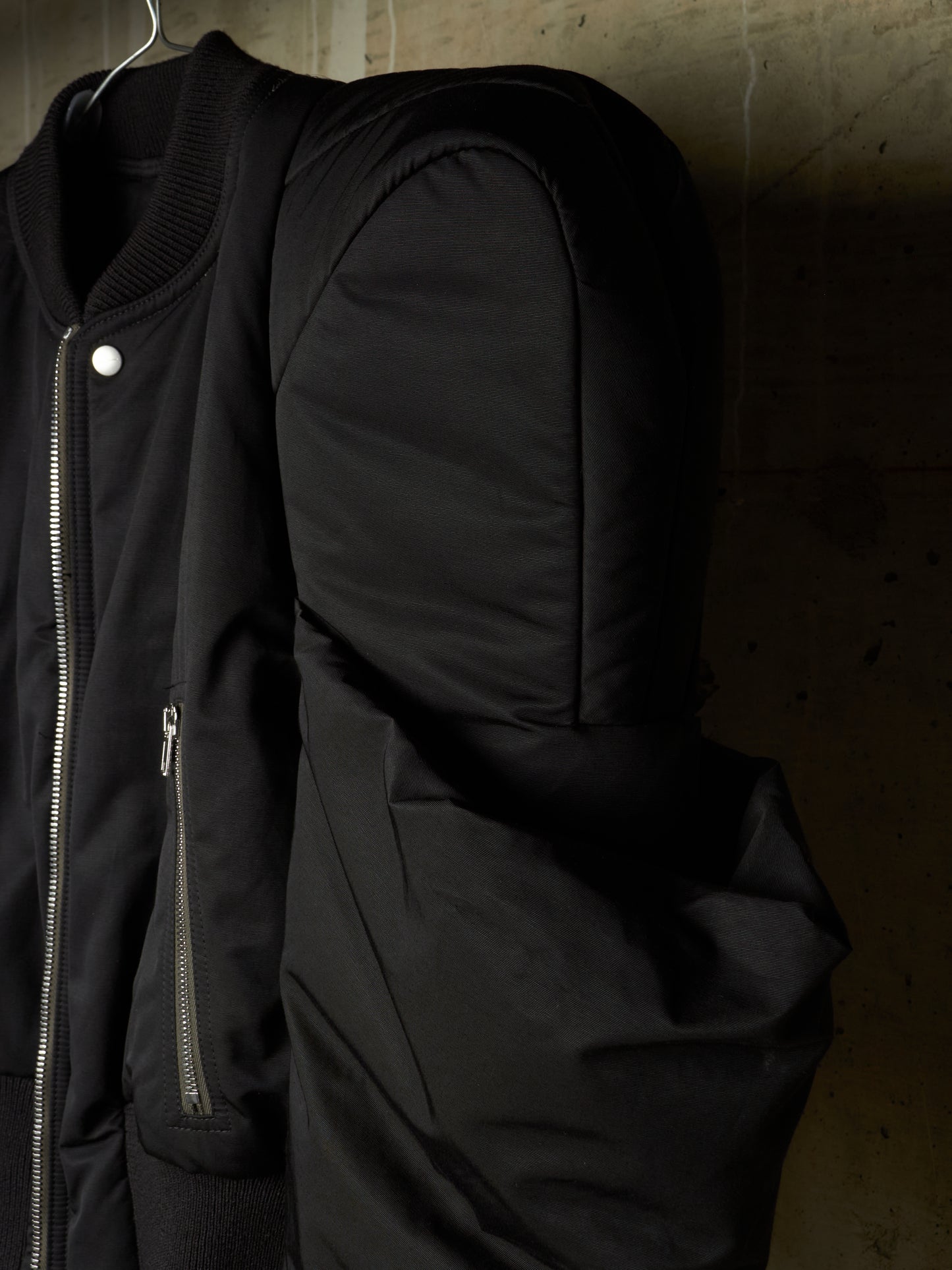 A close-up of a black bomber jacket hanging on a hook against a textured concrete wall, showing the ribbed collar, silver zipper, and side pocket.