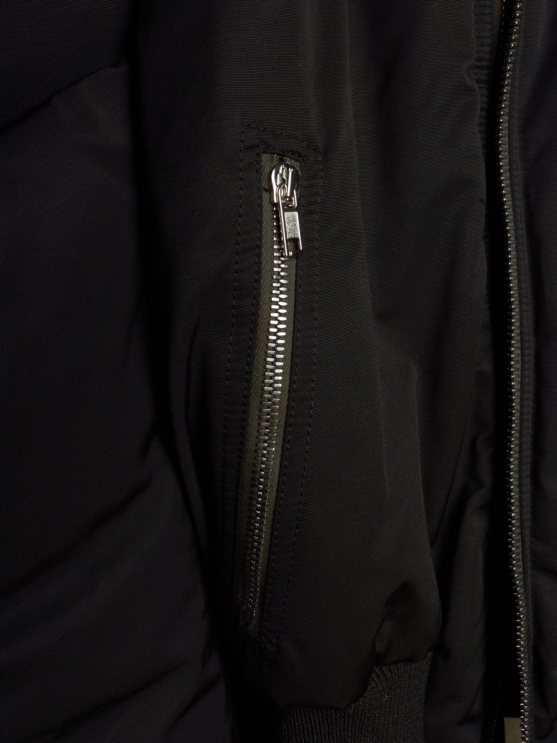 Close-up of a black puffer jacket featuring a vertical zippered pocket with a silver zipper pull and a main front zipper.