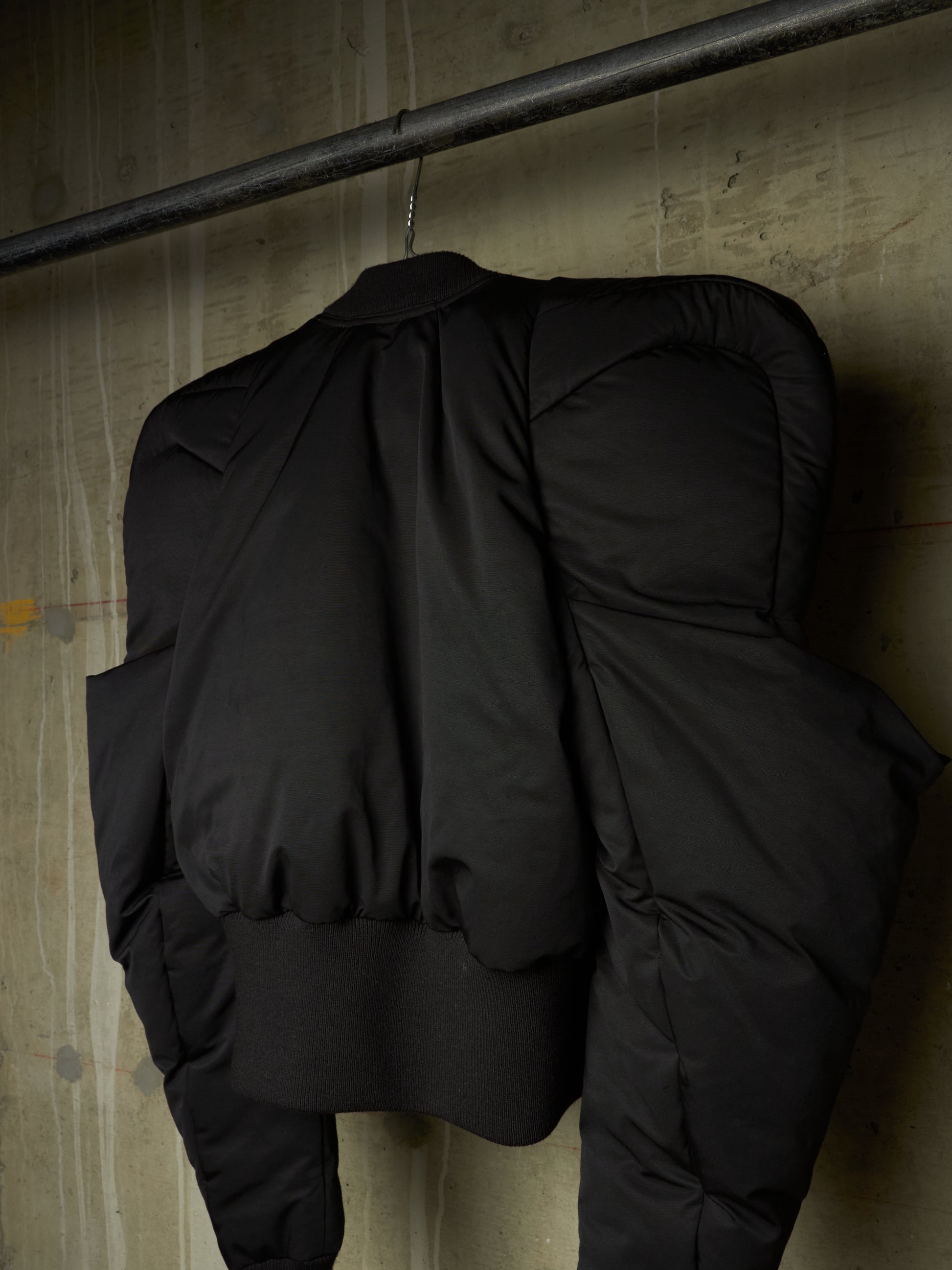 A black puffer jacket hanging from a metal rod against a textured concrete wall, showing the back view with padded shoulders and a ribbed hem.
