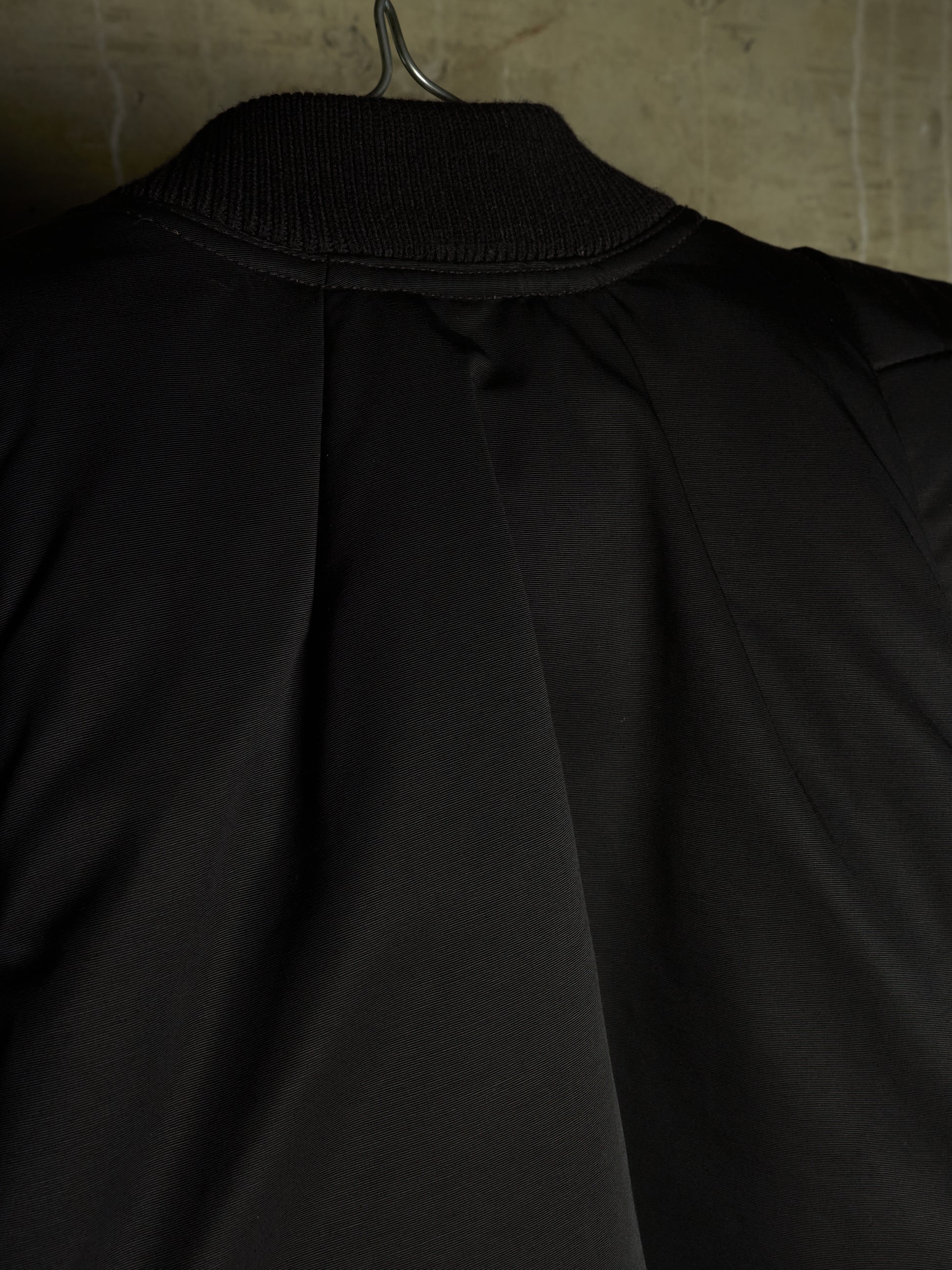 Close-up of the back of a black jacket hanging on a metal hanger against a textured beige wall.