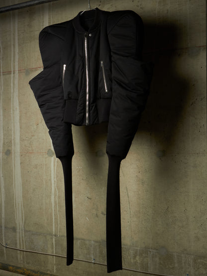 A black puffer jacket with dramatic shoulder padding, a front zipper, and long dangling tie straps, hanging against a textured concrete wall.