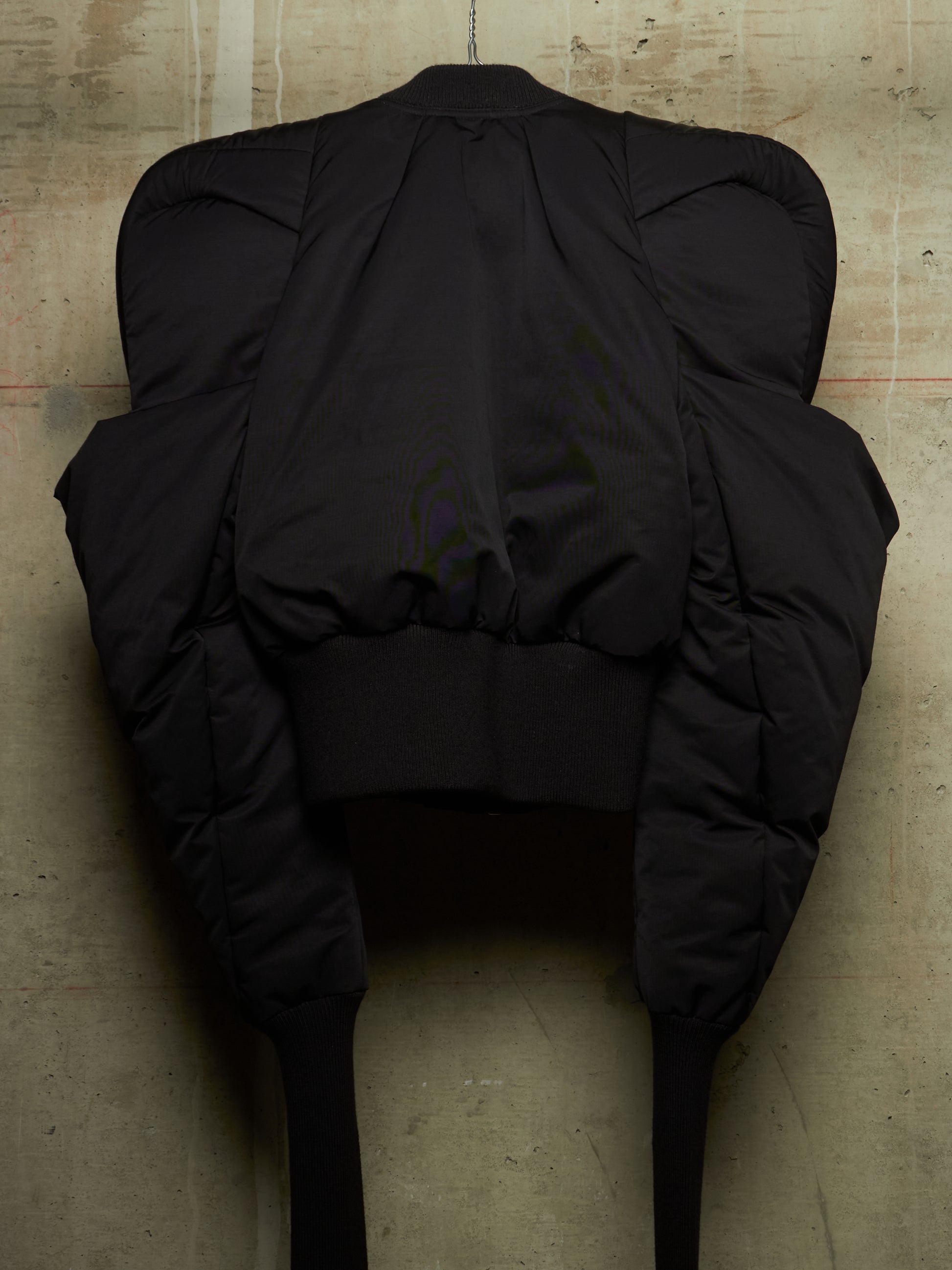 A black puffer jacket with dramatic, oversized padded shoulders and long legs, shown from the back against a textured concrete wall.