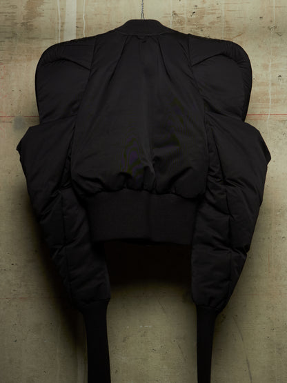 A black puffer jacket with dramatic, oversized padded shoulders and long legs, shown from the back against a textured concrete wall.