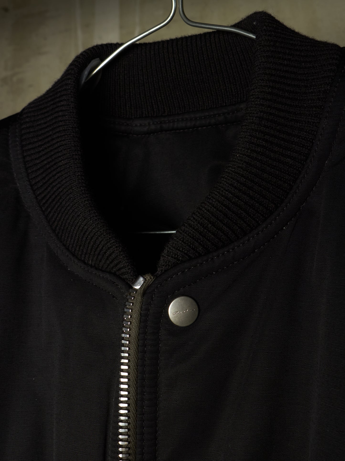 Close-up of a black zip-front jacket hanging on a metal hanger, showing a ribbed knit collar and a silver snap button.