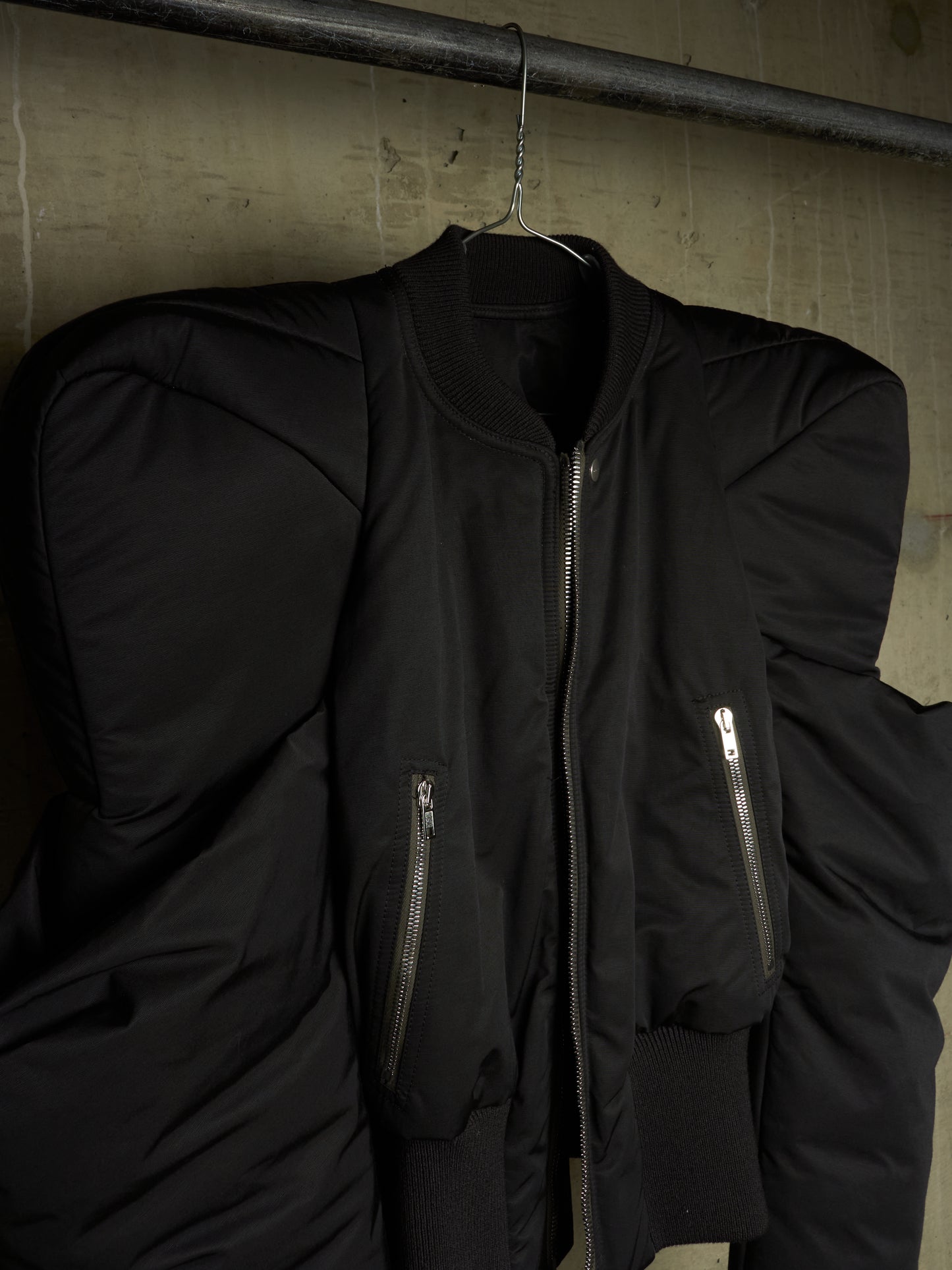 A black oversized puffer bomber jacket with dramatic shoulder padding, hanging on a metal hanger against a textured concrete wall.