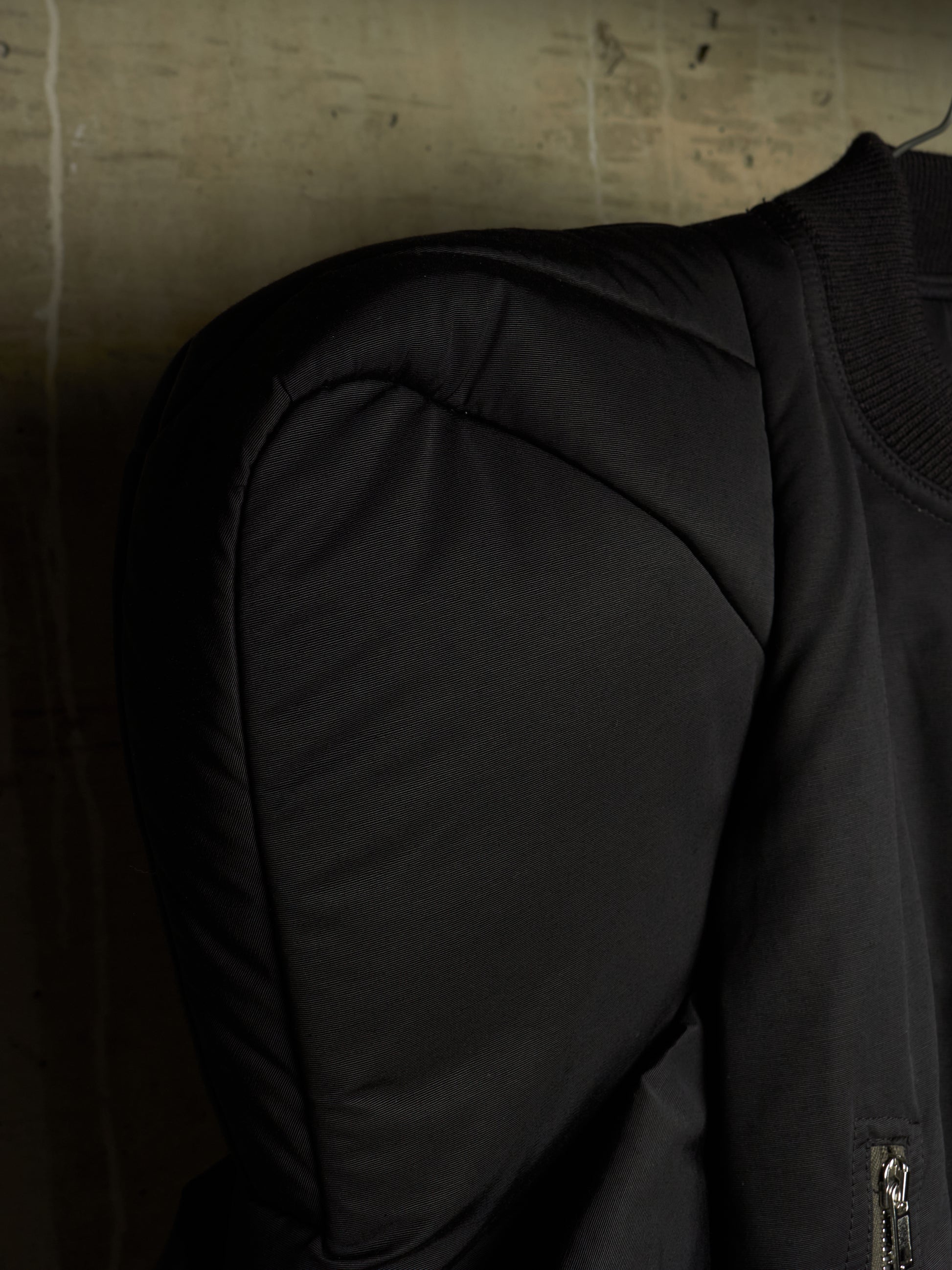 Close-up of a black puffer jacket sleeve and shoulder, showing padded seam details and a ribbed collar, set against a dark textured background.