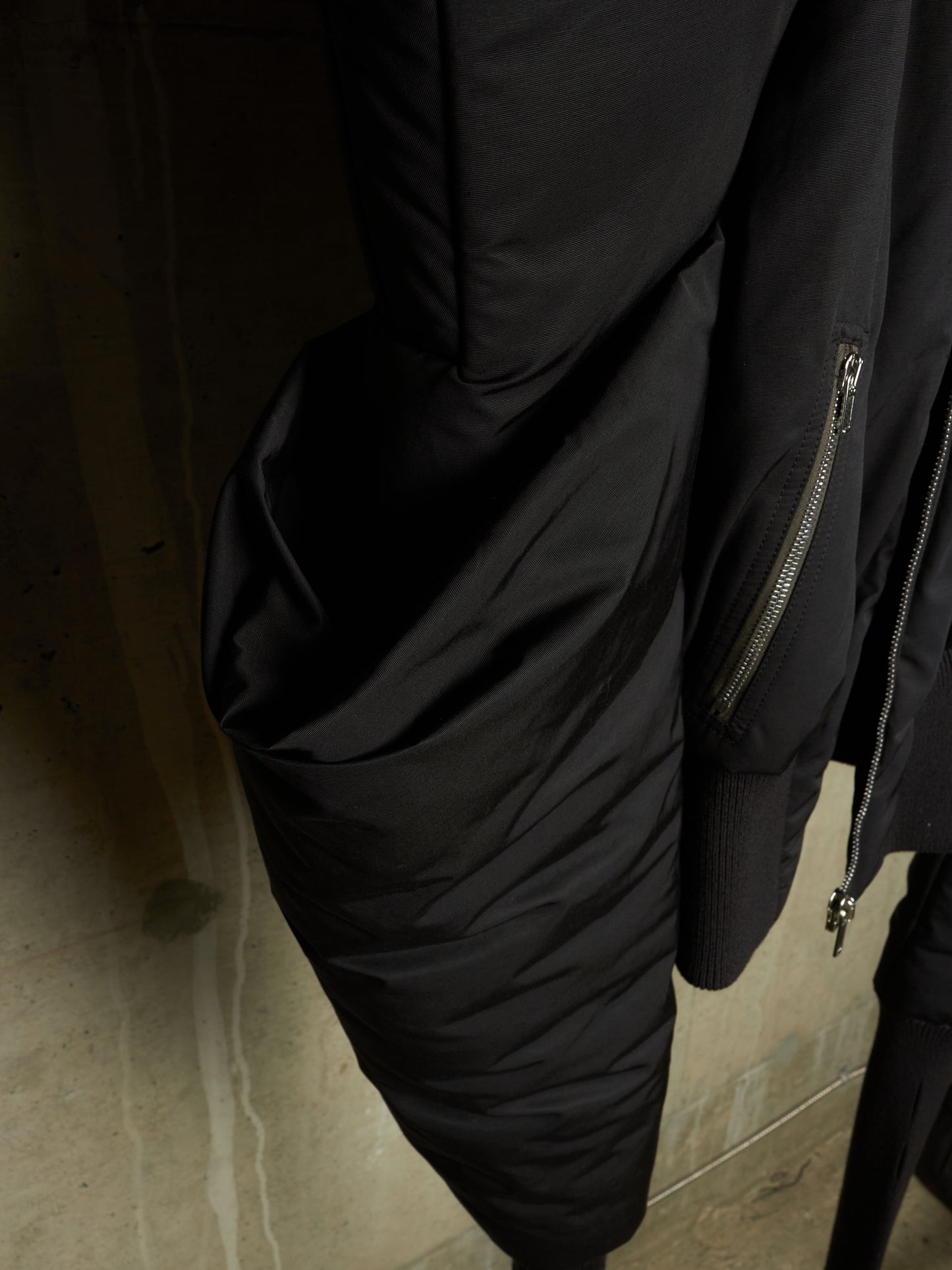 Close-up of a black puffer jacket with quilted sections, ribbed cuffs, and silver zipper details, hanging against a dark textured wall.