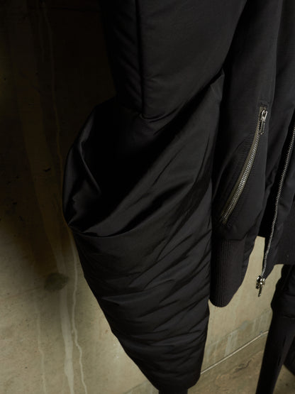 Close-up of a black puffer jacket with quilted sections, ribbed cuffs, and silver zipper details, hanging against a dark textured wall.