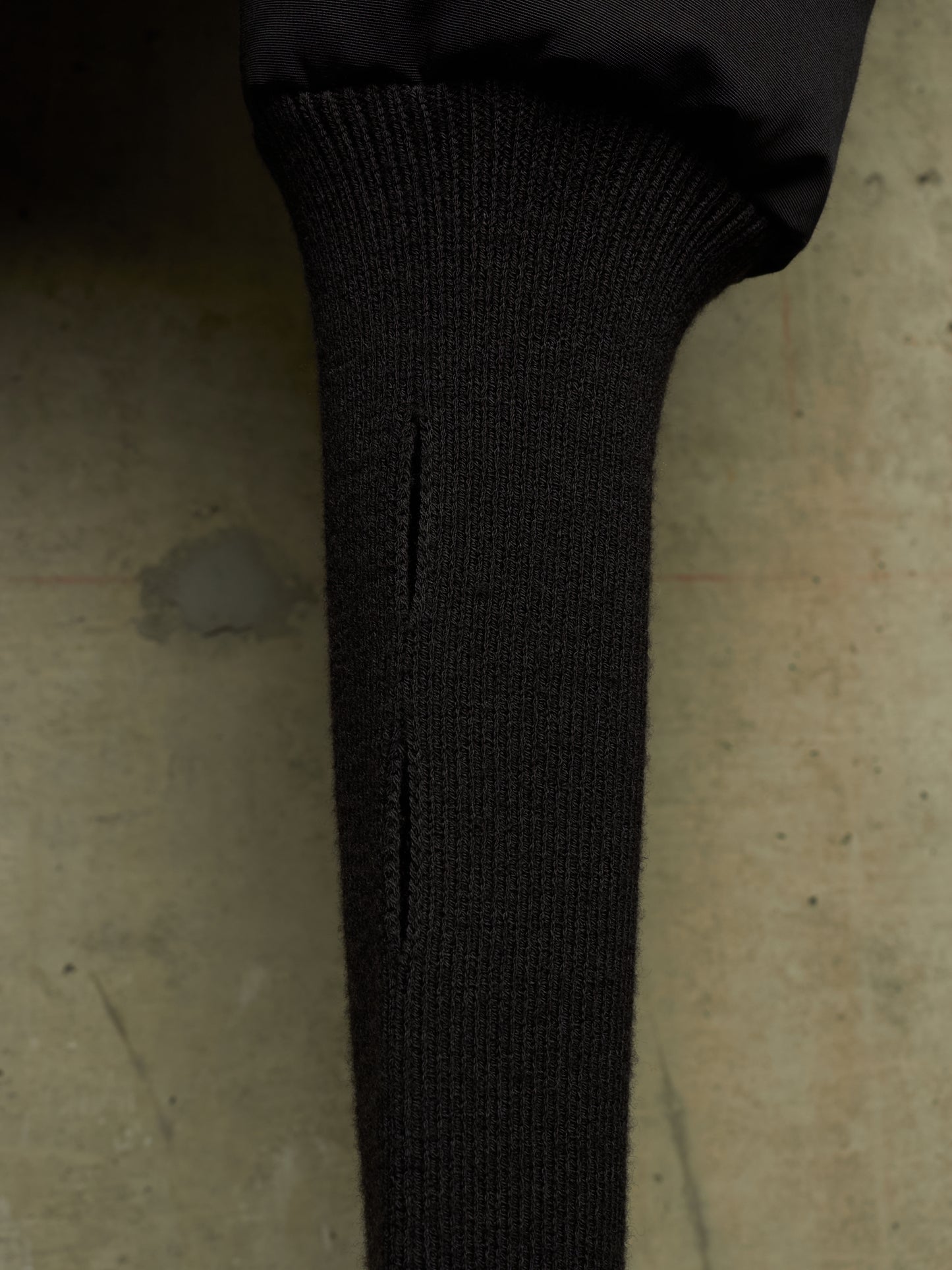 Close-up of a black ribbed knit sleeve or cuff with a vertical slit, attached to a black puffer jacket, set against a textured concrete background.