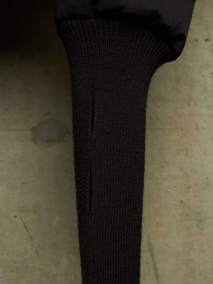 Close-up of a black ribbed knit sleeve or cuff with a vertical slit, attached to a black puffer jacket, set against a textured concrete background.