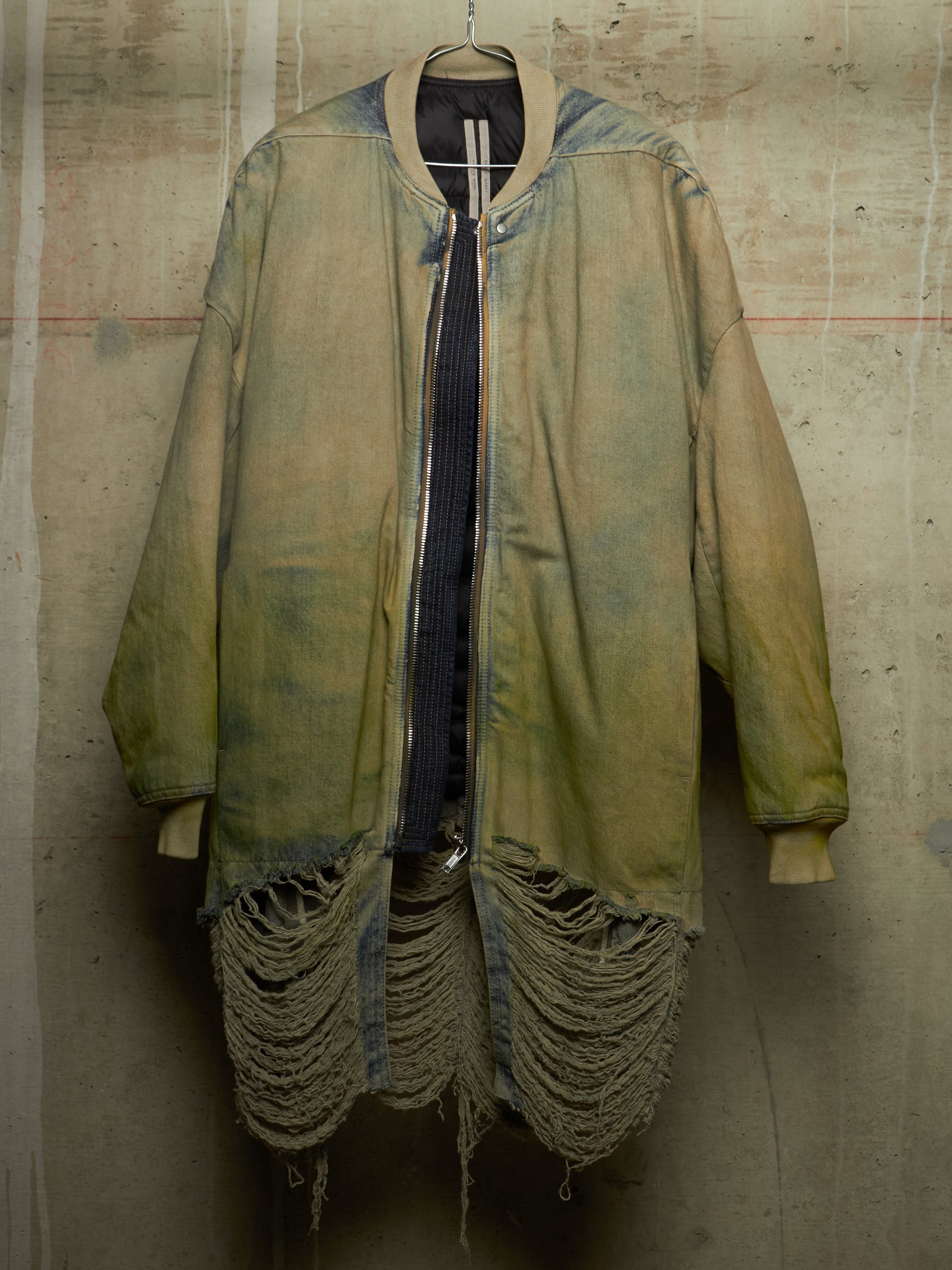 A distressed oversized bomber-style jacket in faded olive green and tan, hanging against a textured concrete wall. The jacket features heavy fraying and tattered detailing along the hem and cuffs, with a dark ribbed shirt underneath.
