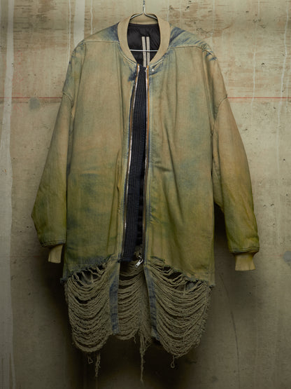 A distressed oversized bomber-style jacket in faded olive green and tan, hanging against a textured concrete wall. The jacket features heavy fraying and tattered detailing along the hem and cuffs, with a dark ribbed shirt underneath.