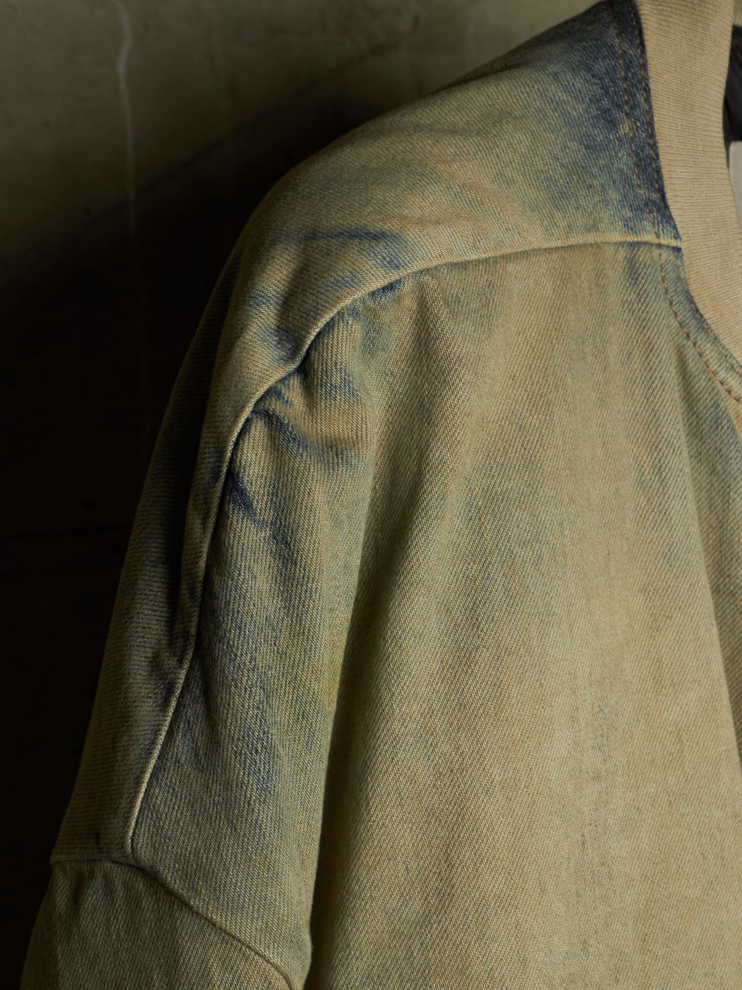 A close-up detail shot of a washed, distressed denim jacket showing the shoulder seam, sleeve, and collar area in a muted beige and blue-gray colorway.