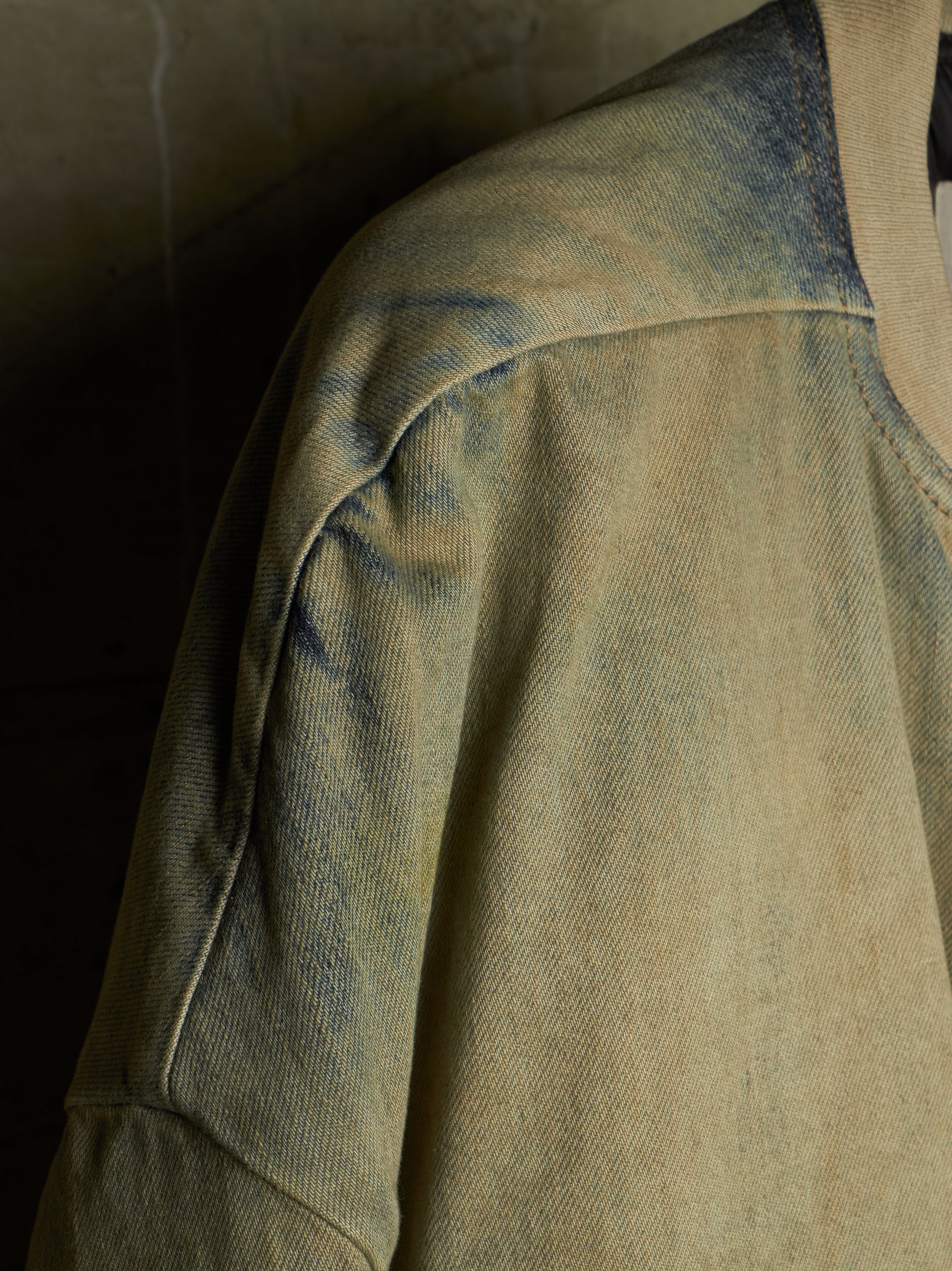 A close-up detail shot of a washed, distressed denim jacket showing the shoulder seam, sleeve, and collar area in a muted beige and blue-gray colorway.