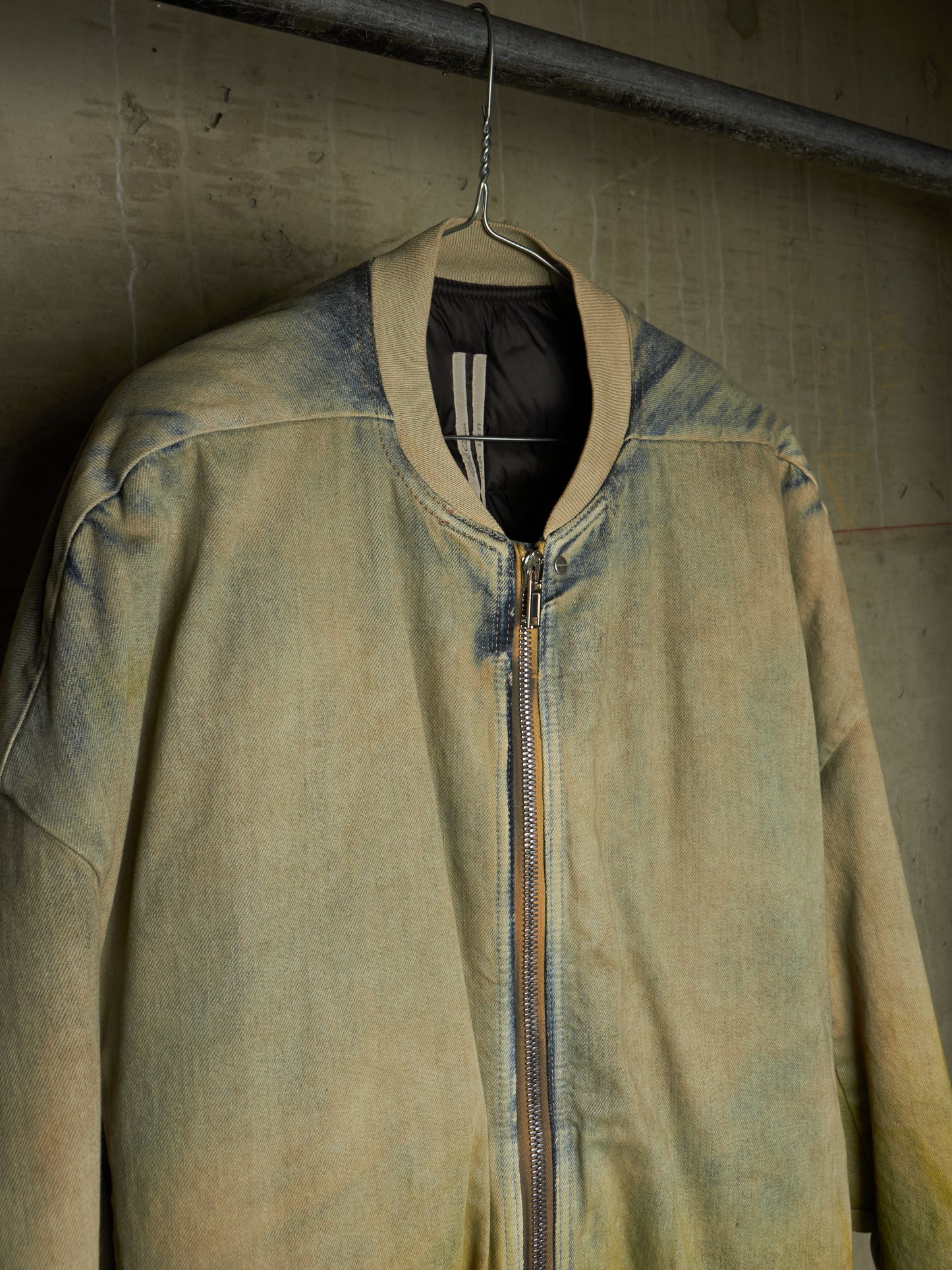 A faded olive green bomber jacket with a distressed, washed texture, hanging on a wire hanger against a textured concrete wall.