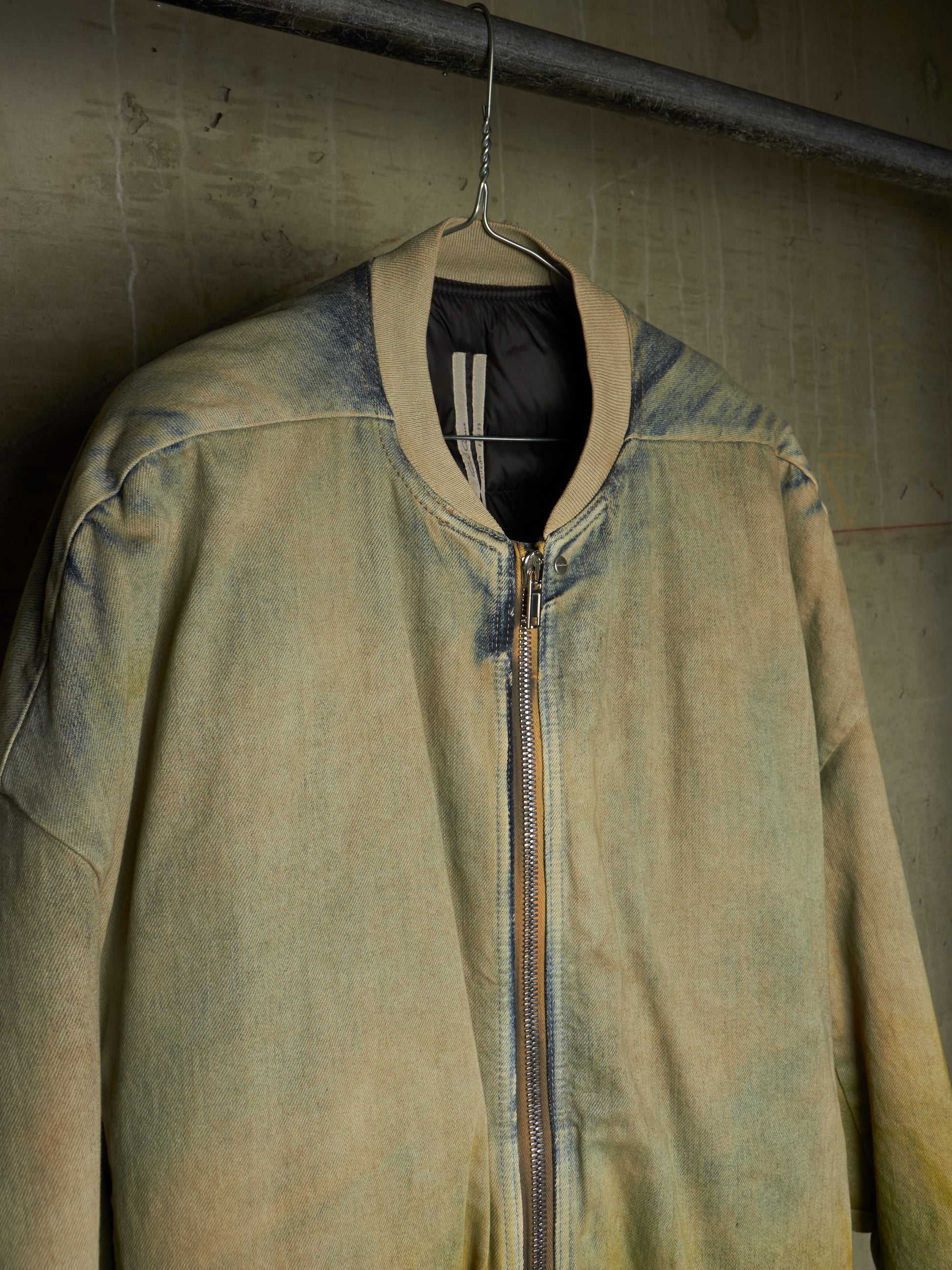 A faded olive green bomber jacket with a distressed, washed texture, hanging on a wire hanger against a textured concrete wall.