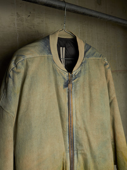 A faded olive green bomber jacket with a distressed, washed texture, hanging on a wire hanger against a textured concrete wall.