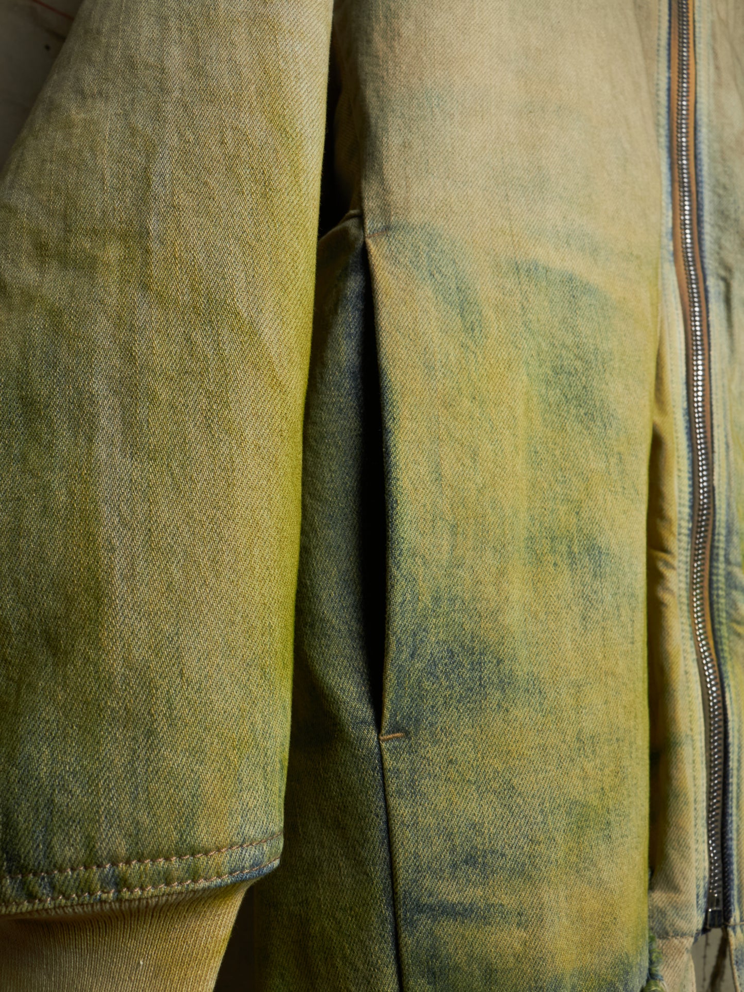 Close-up of a distressed olive green and yellow jacket with a silver zipper, showing detailed fabric texture and seams.