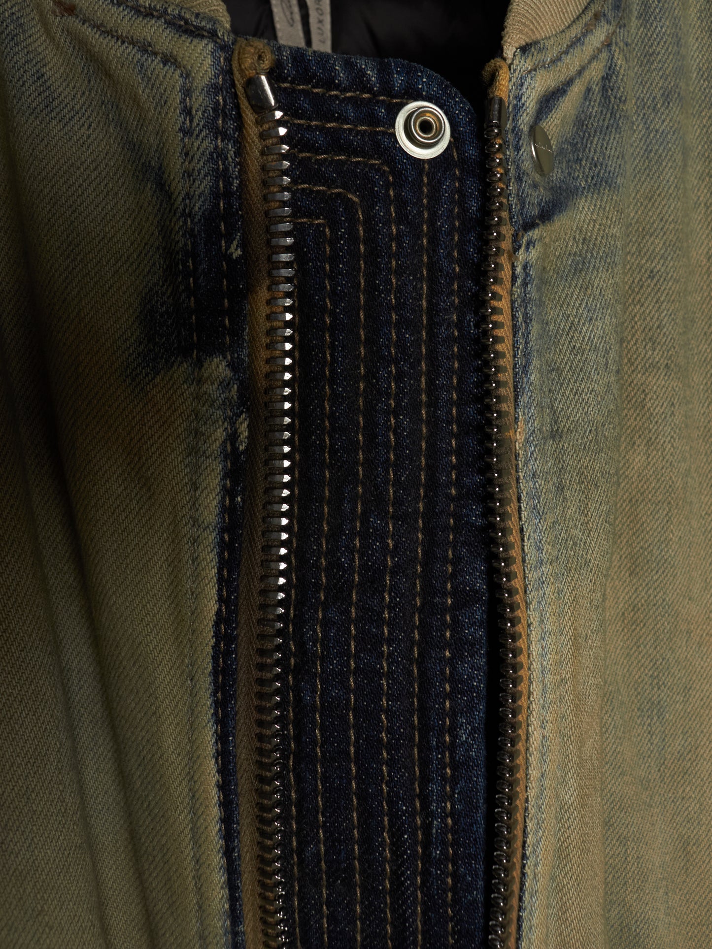 Close-up detail of a jacket's front closure, showing a dark navy denim placket with contrast gold stitching, a silver metal zipper, and a snap button, set against a faded olive green denim fabric.