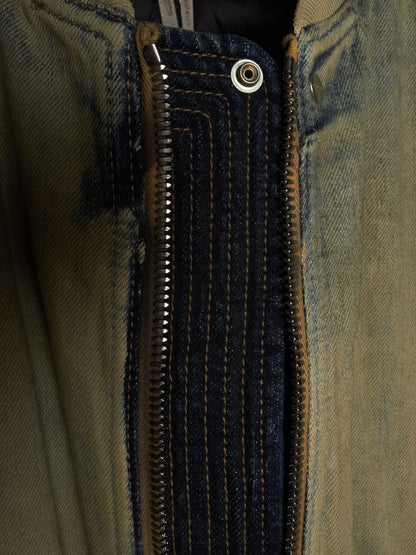 Close-up detail of a jacket's front closure, showing a dark navy denim placket with contrast gold stitching, a silver metal zipper, and a snap button, set against a faded olive green denim fabric.