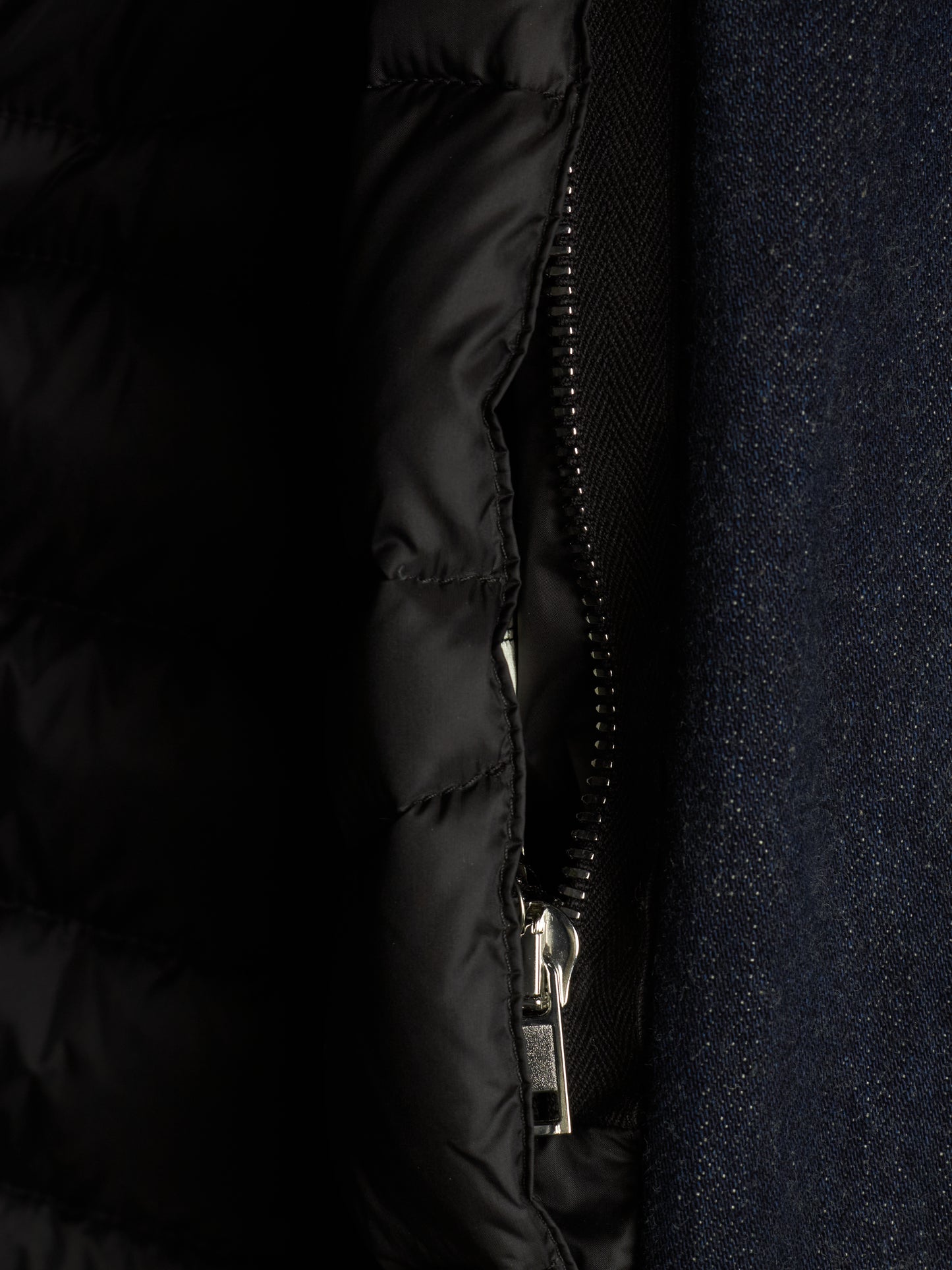 Close-up detail of a black quilted puffer jacket with a silver zipper, shown next to a dark navy textured fabric.